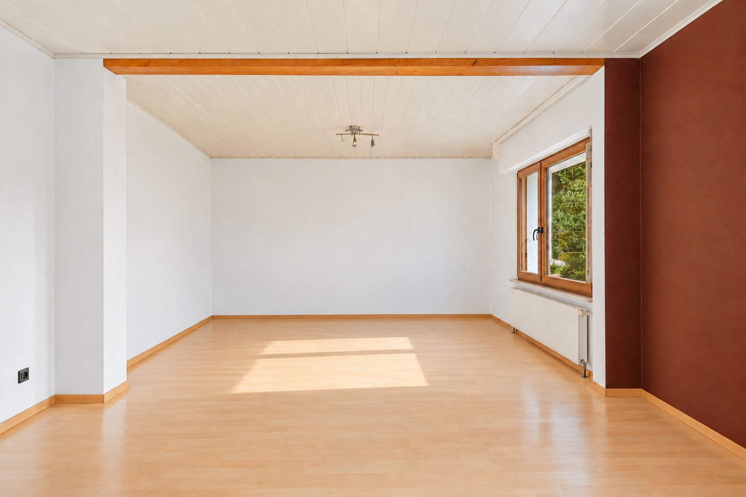 Leerer Raum mit Holzboden, weißer Decke und großem Fenster an der rechten Wand.