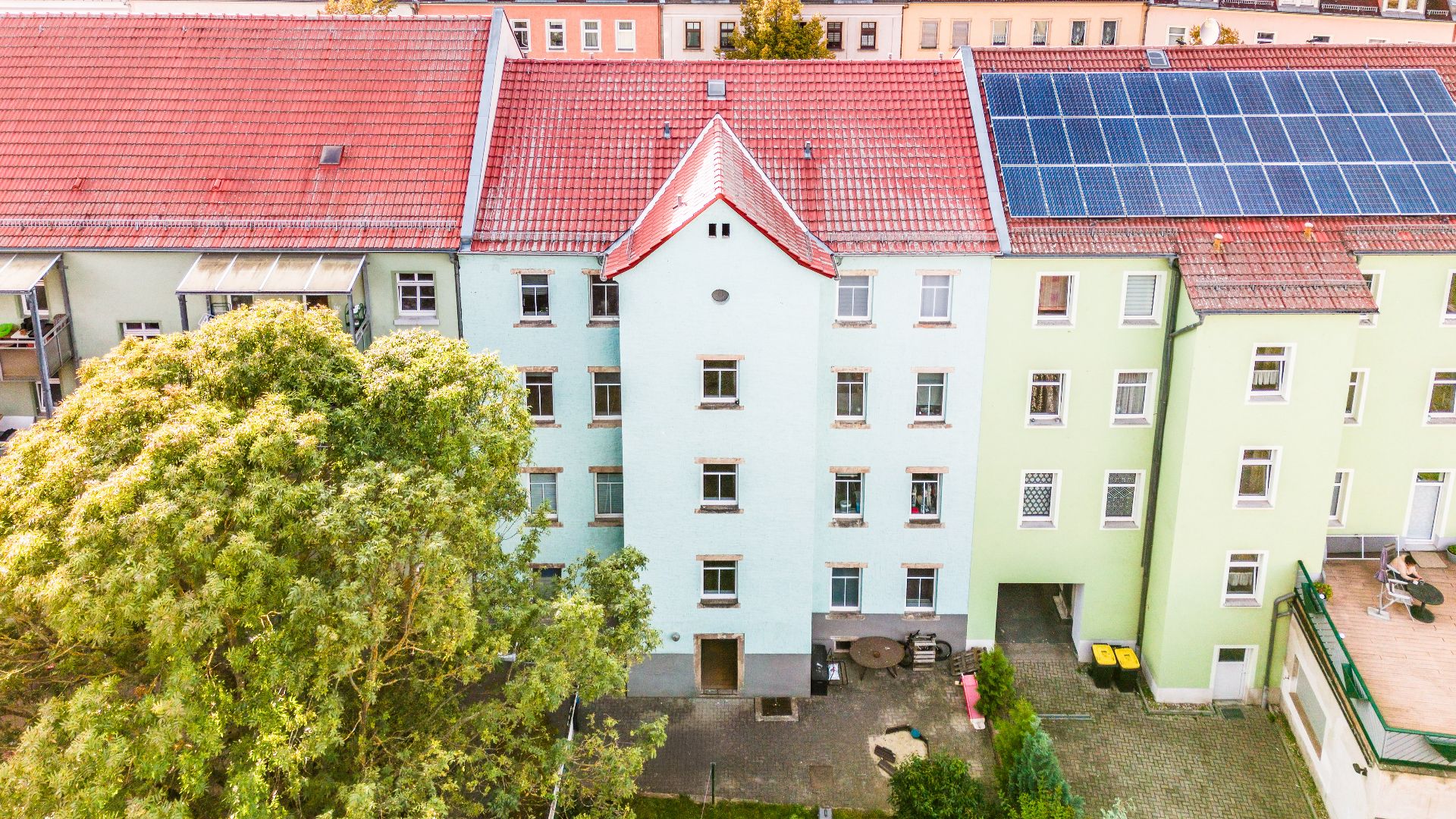 Mehrstöckiges Wohnhaus mit rotem Dach, Solarpaneelen und Bäumen im Vordergrund.