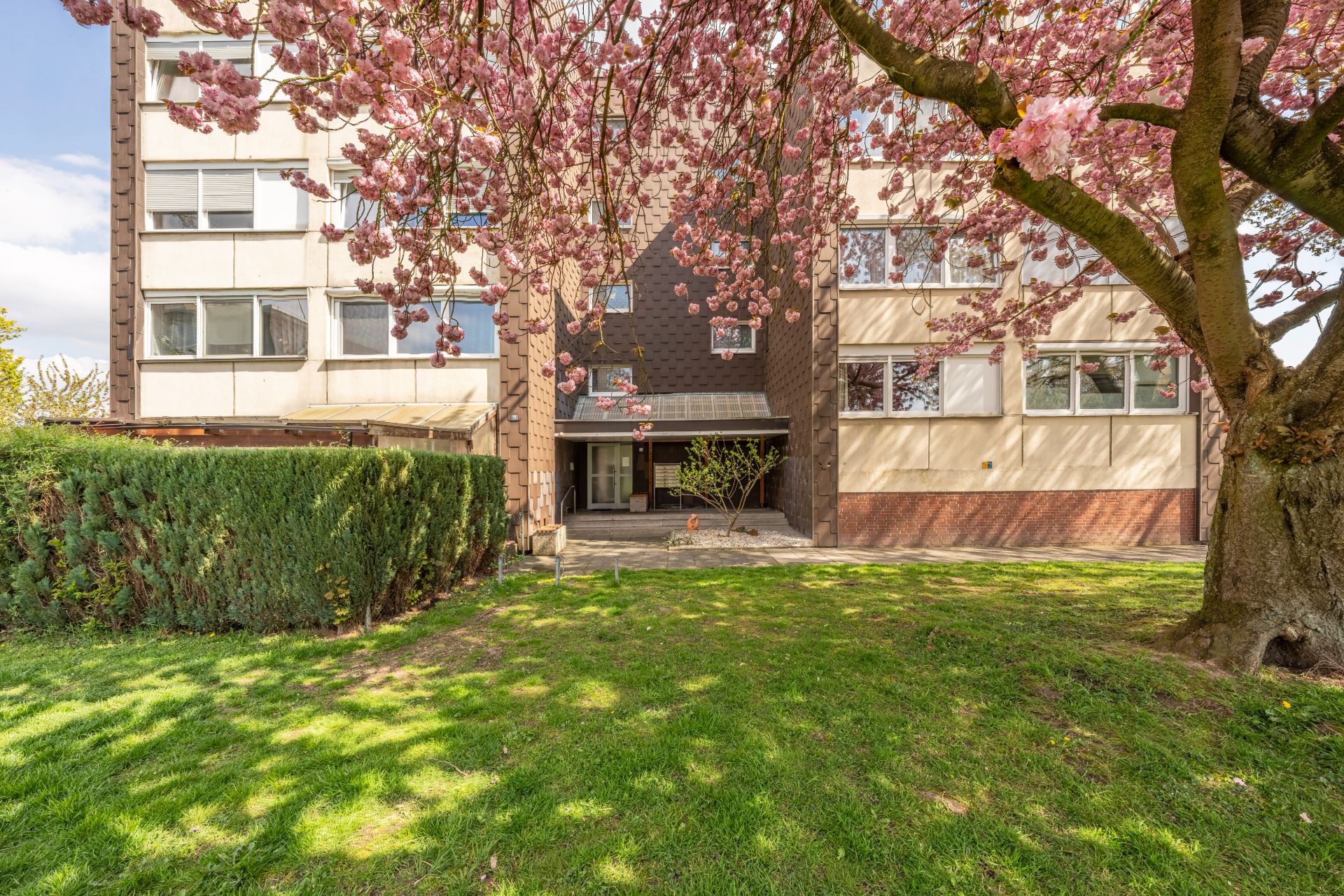 Mehrfamilienhaus mit blühendem Baum und grünem Rasen im Vordergrund.
