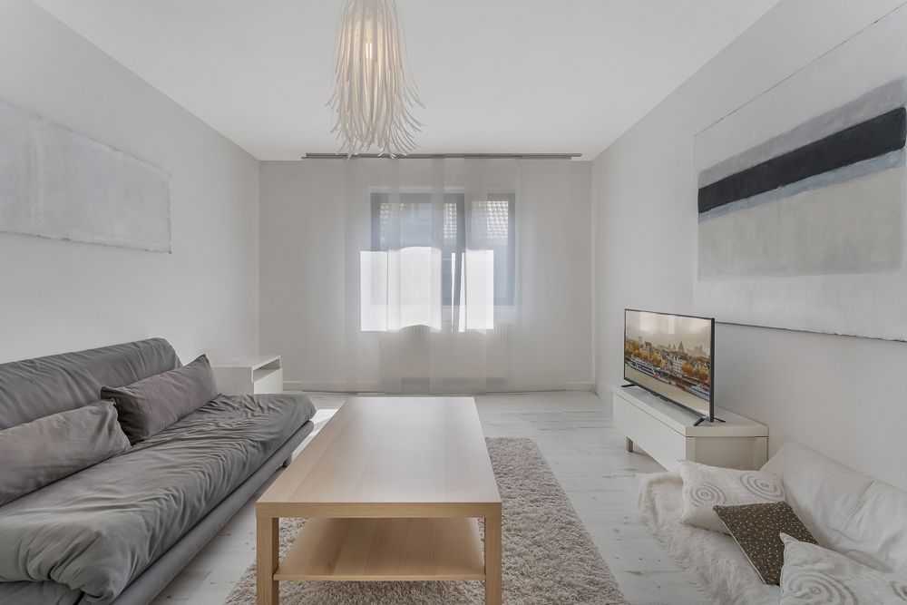 Helles Wohnzimmer mit grauem Sofa, Holztisch, Fernseher und großen Fenstern mit Vorhängen.