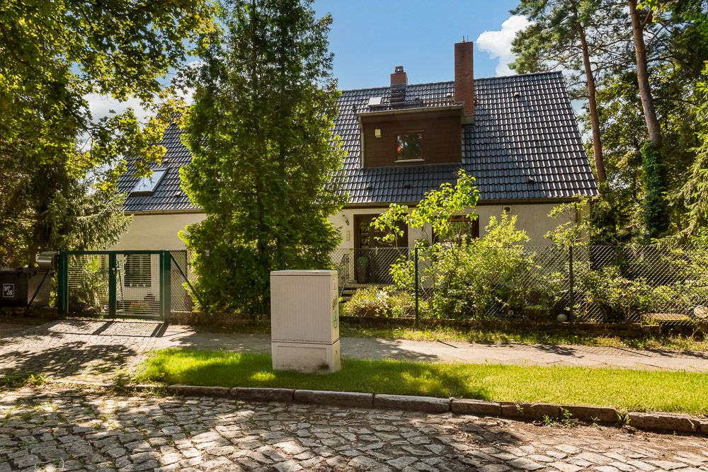 Haus mit dunklem Dach und Garten, von Bäumen und Zaun umgeben, davor steht ein Verteilerkasten.