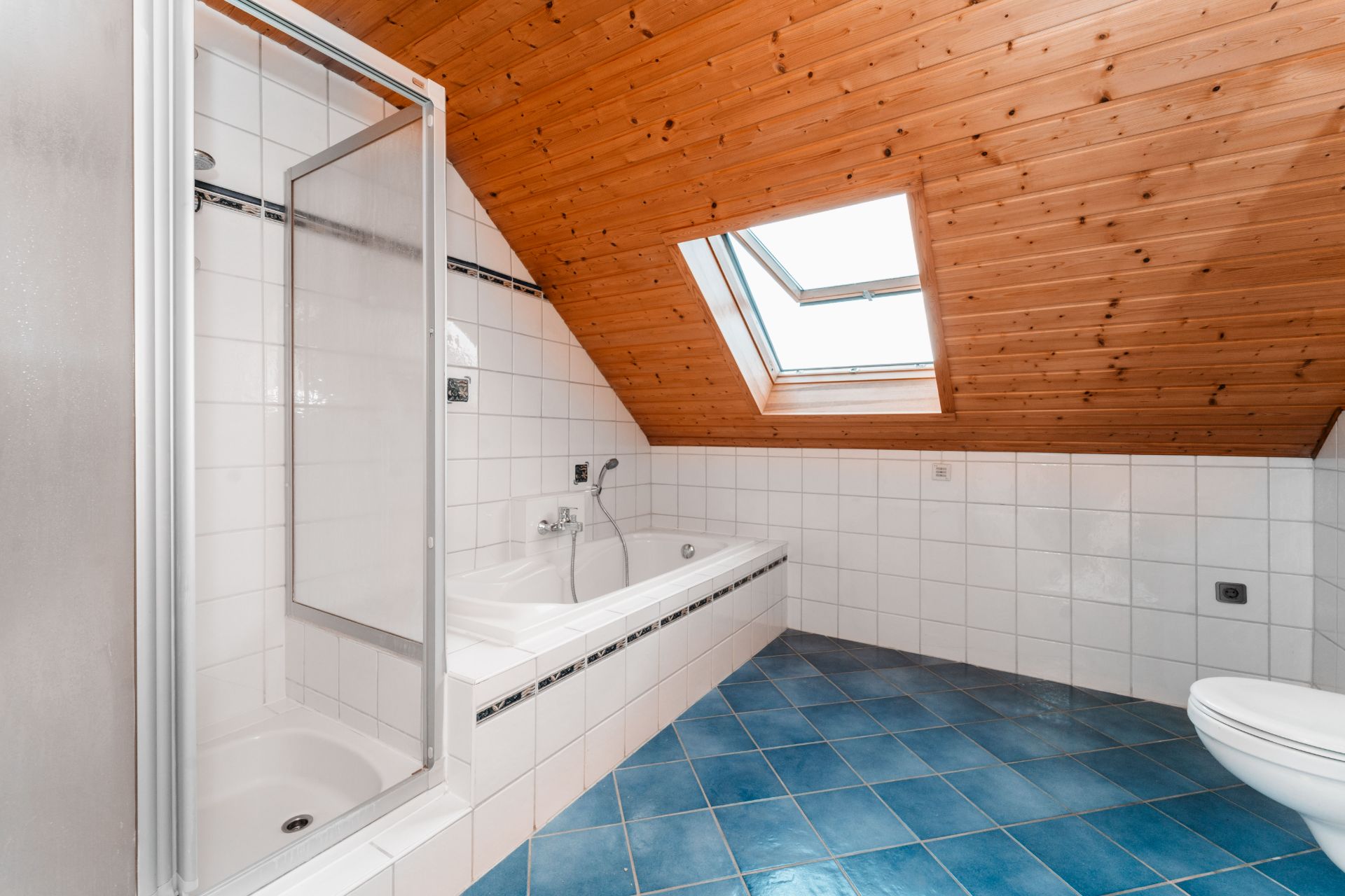 Badezimmer mit Dusche, Badewanne, blauen Fliesen und Dachfenster unter Holzdecke.