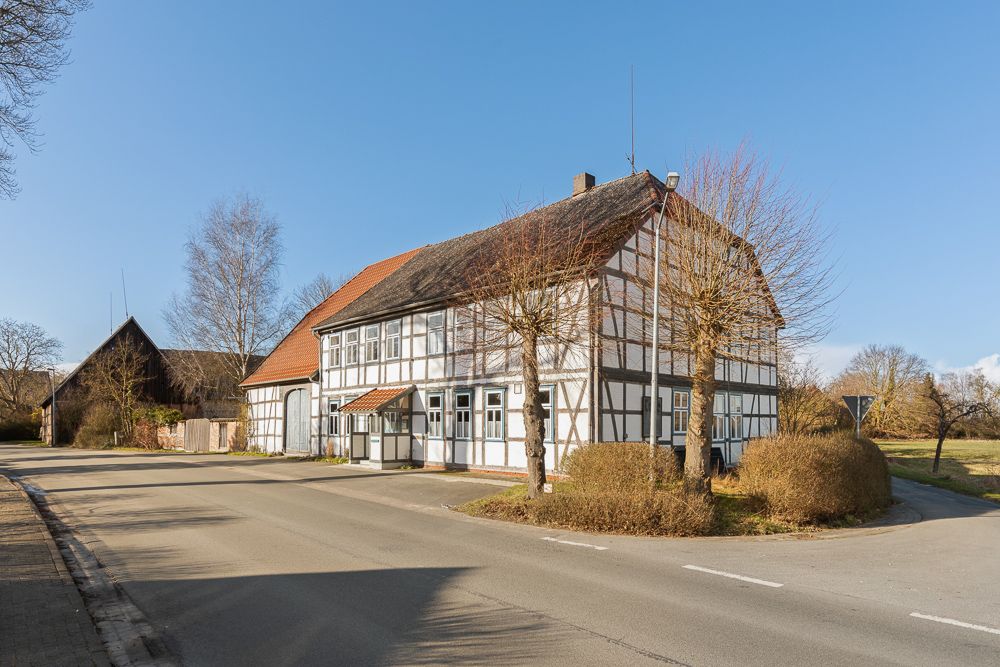 Fachwerkhaus mit rotem Dach an einer Straßenecke, umgeben von kahlen Bäumen und Büschen.