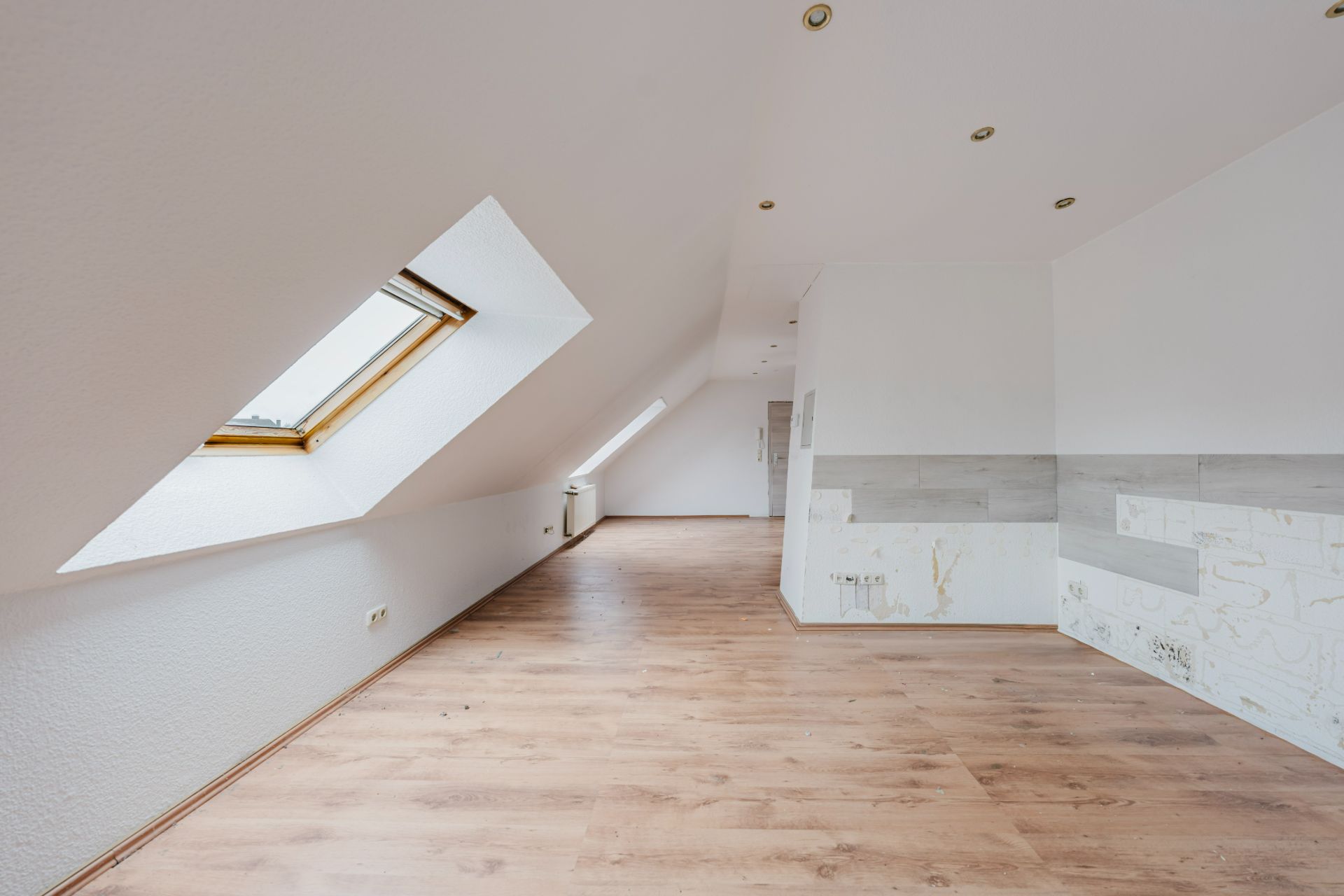 Heller Dachgeschossraum mit schrägen Fenstern, Holzfußboden und weißen Wänden.
