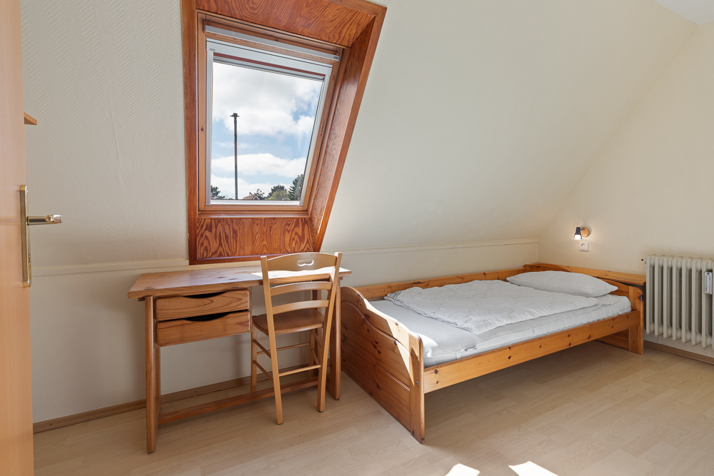 Helles Zimmer mit Einzelbett, Schreibtisch, Stuhl und Dachfenster.