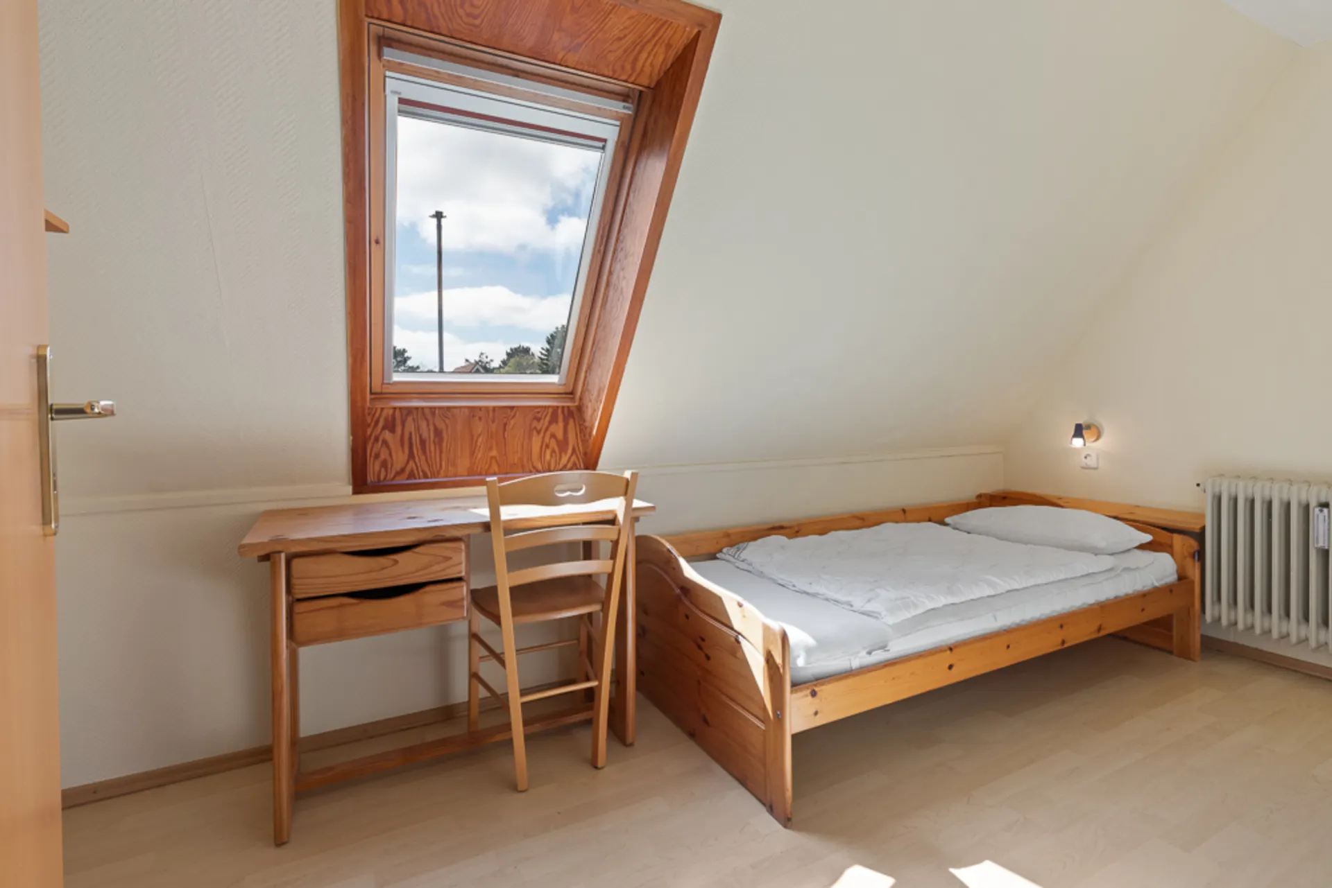 Helles Zimmer mit Einzelbett, Schreibtisch, Stuhl und Dachfenster.