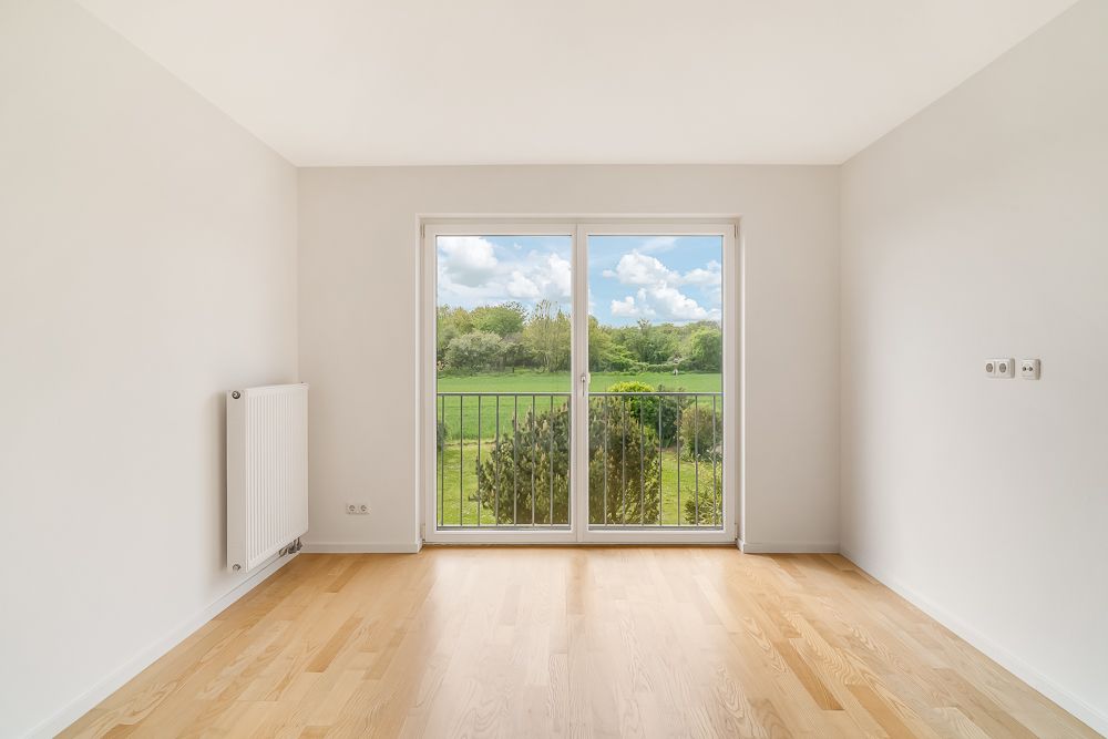 Leeres Zimmer mit Holzfußboden und bodentiefen Fenstern mit Blick ins Grüne.