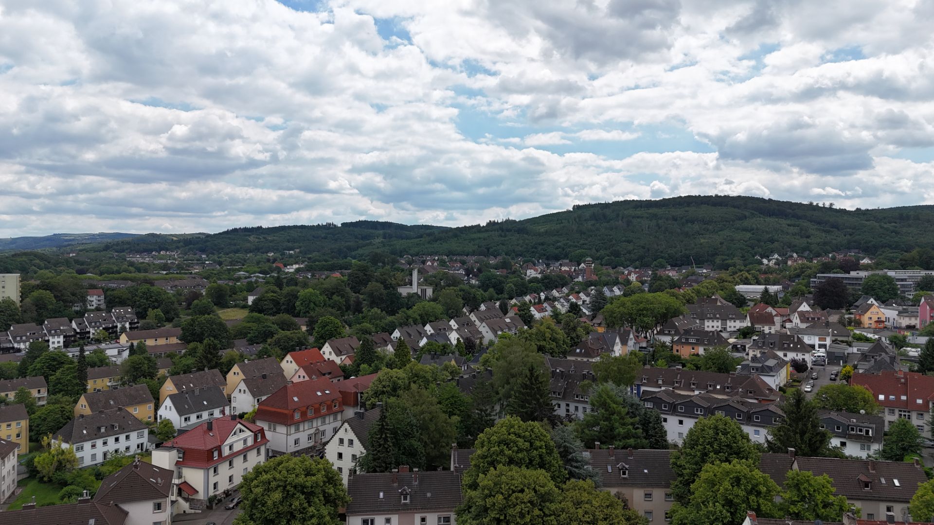 Blick auf Wohnhäuser mit roten und weißen Dächern, viel Grün und bewaldete Hügel im Hintergrund.