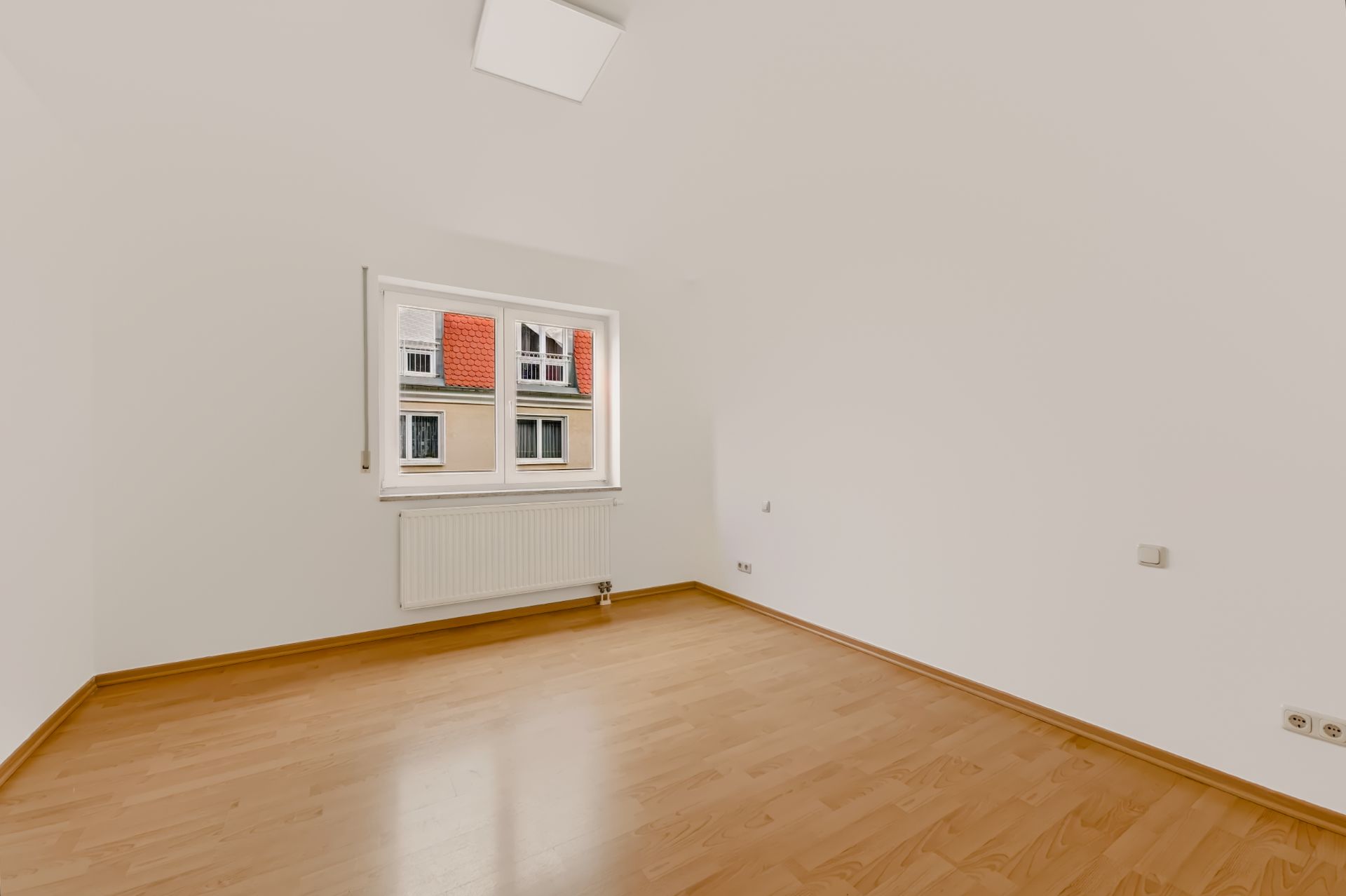Leeres Zimmer mit weißer Wand, Holzfußboden und Fenster mit Blick auf Nachbargebäude.