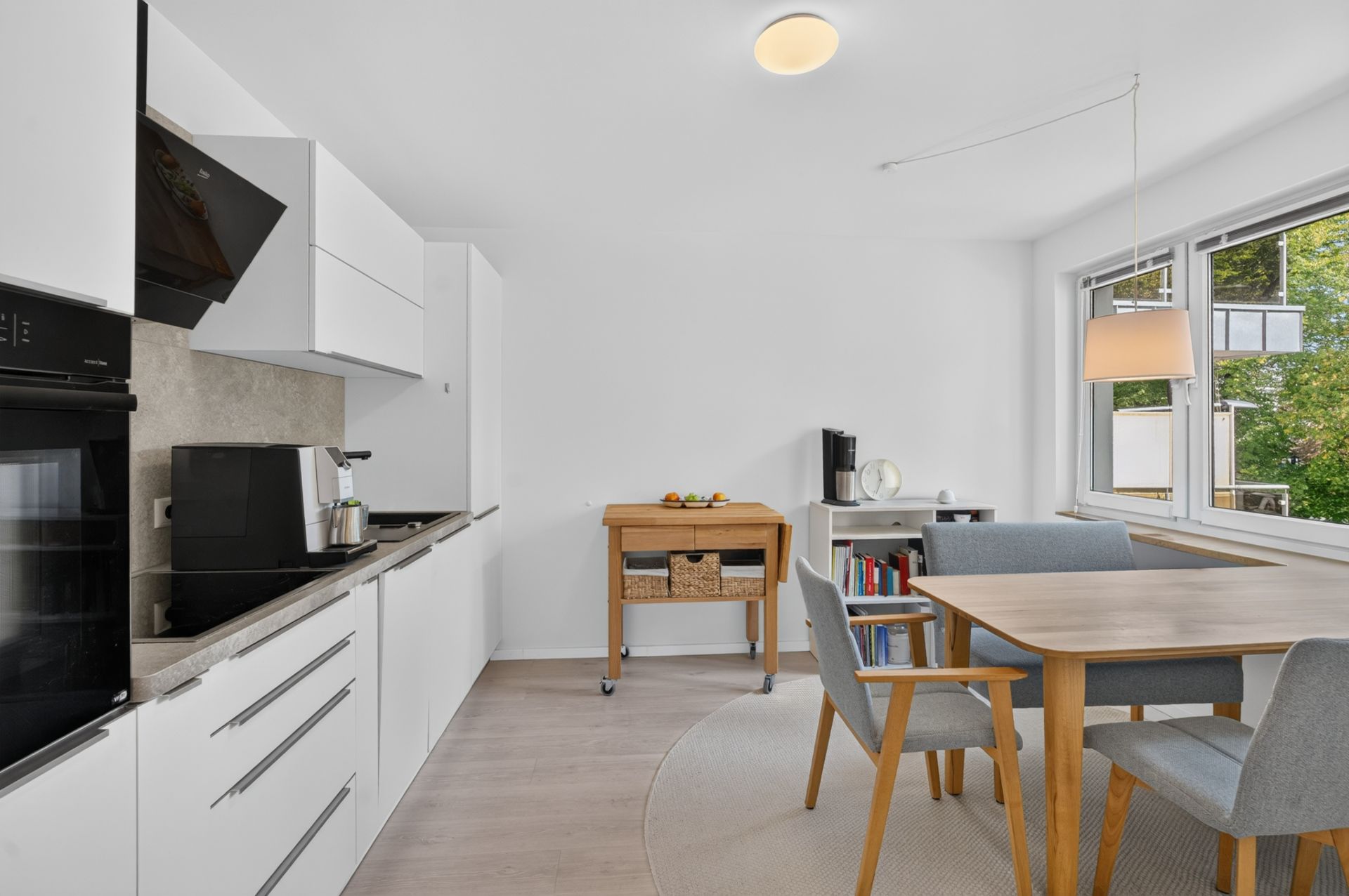 Moderne, helle Küche mit Esstisch und vier Stühlen, großem Fenster und Küchenwagen.