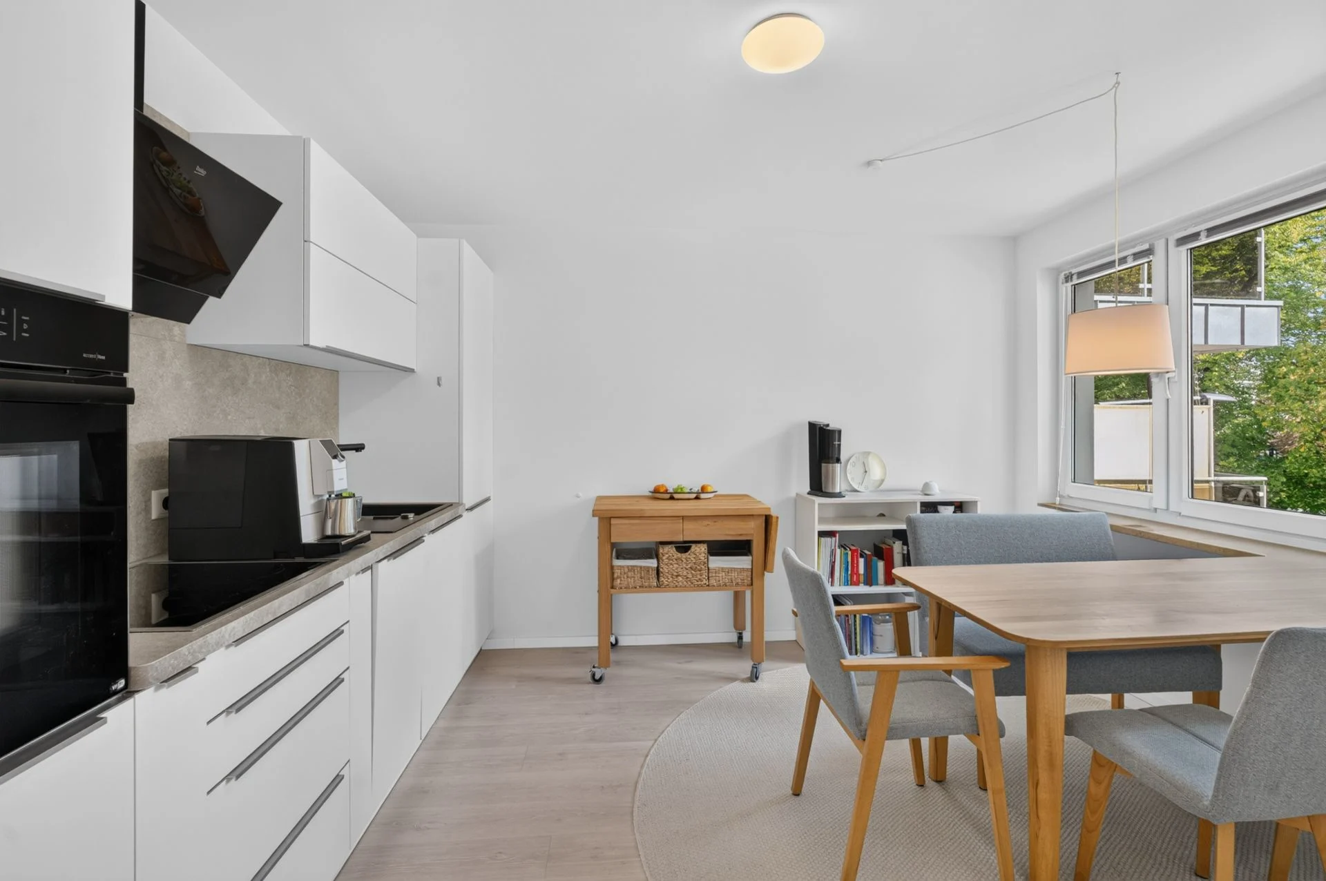 Moderne, helle Küche mit Esstisch und vier Stühlen, großem Fenster und Küchenwagen.