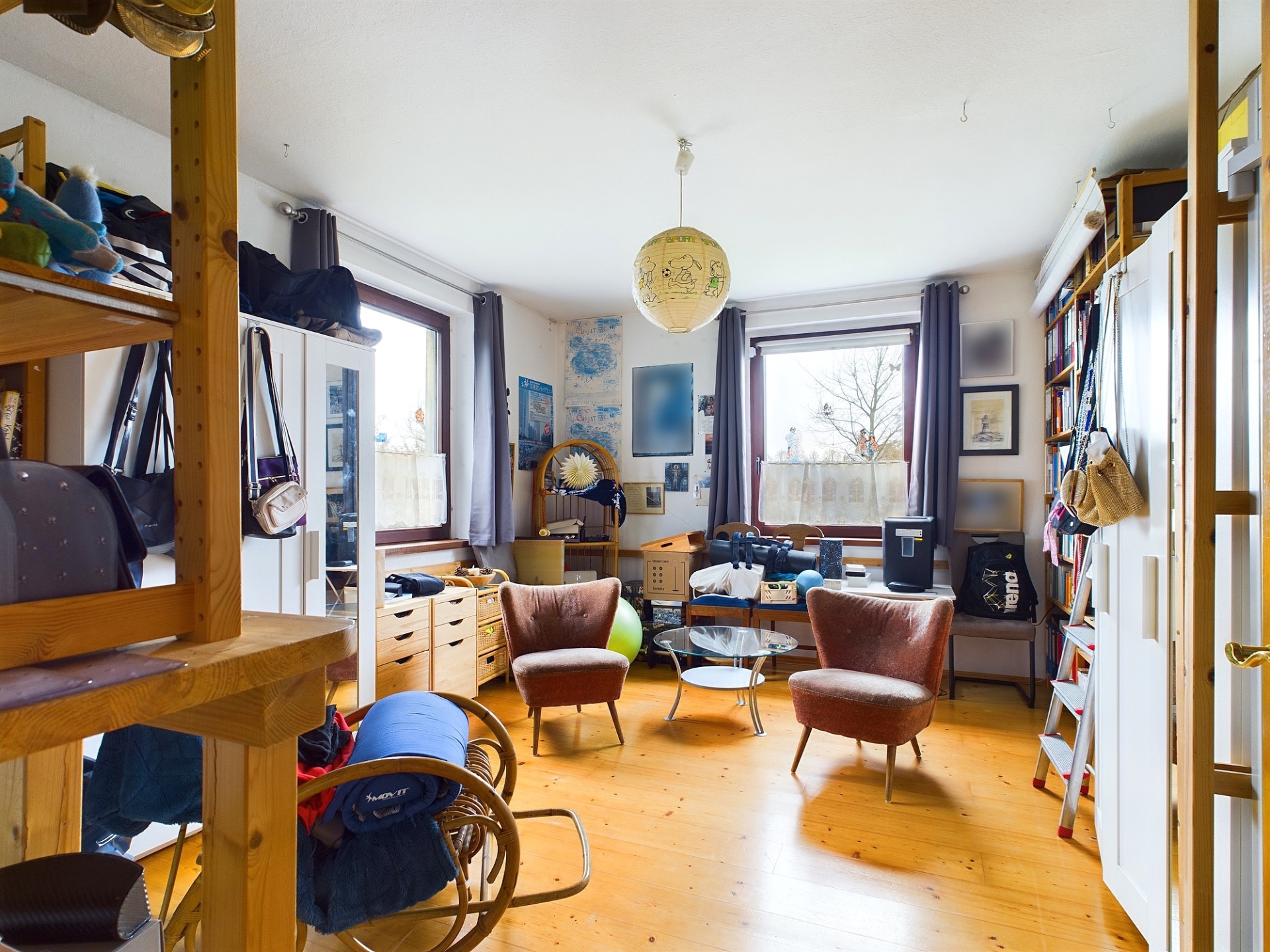 Wohnzimmer mit zwei Sesseln, Bücherregal, Holzboden und zwei großen Fenstern.
