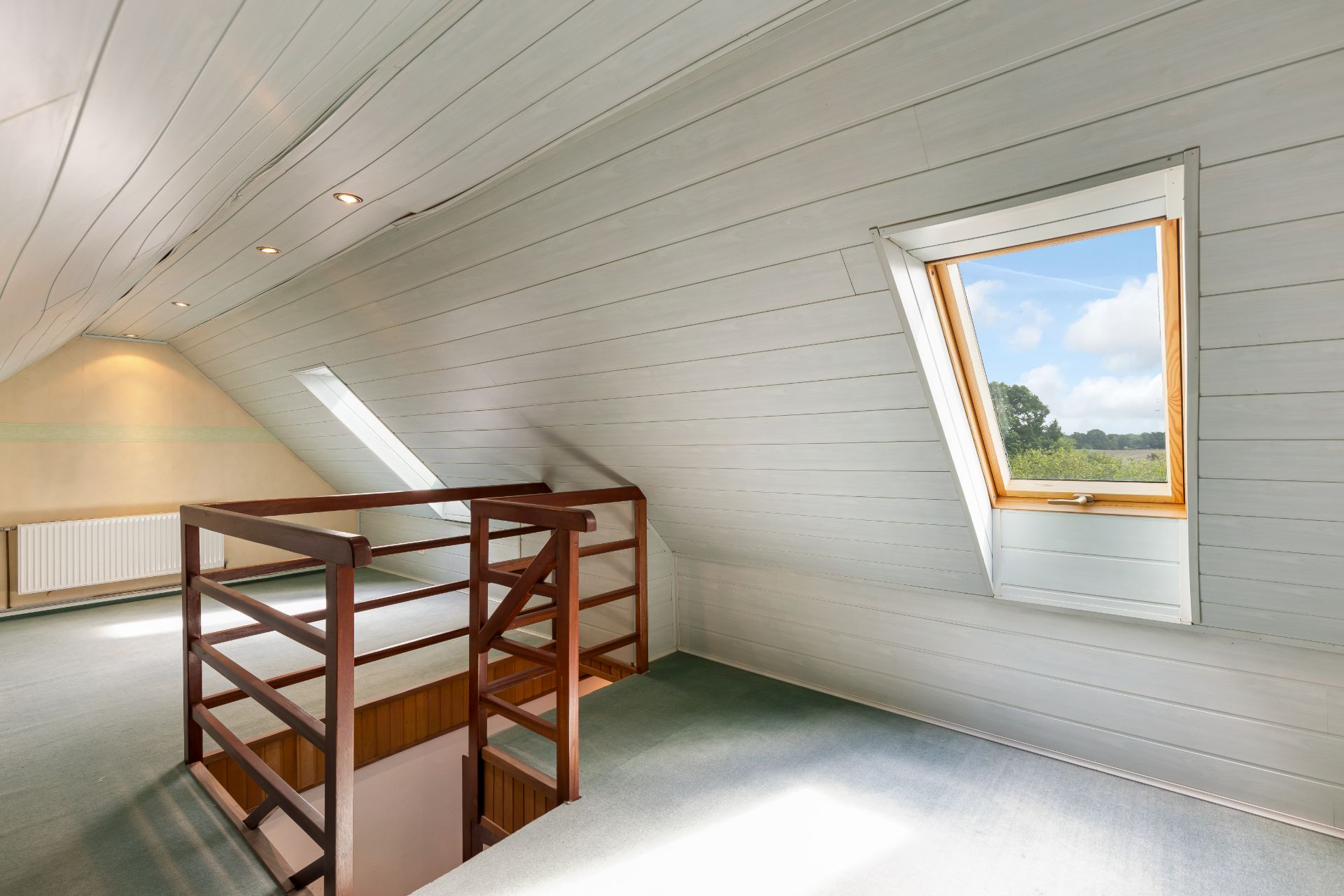 Heller Dachbodenraum mit Teppichboden, Holzgeländer und schrägem Fenster mit Ausblick.