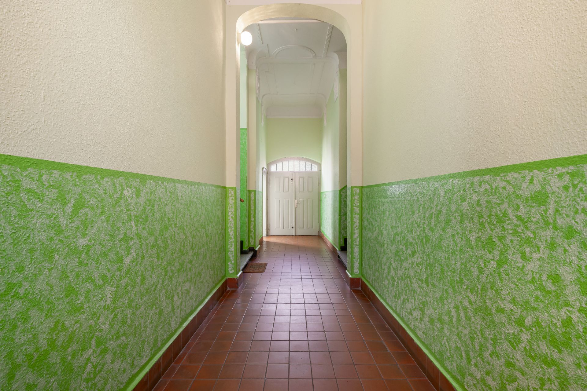 Langer Flur mit grün gestrichenen Wänden und braunen Fliesen, am Ende eine weiße Tür.