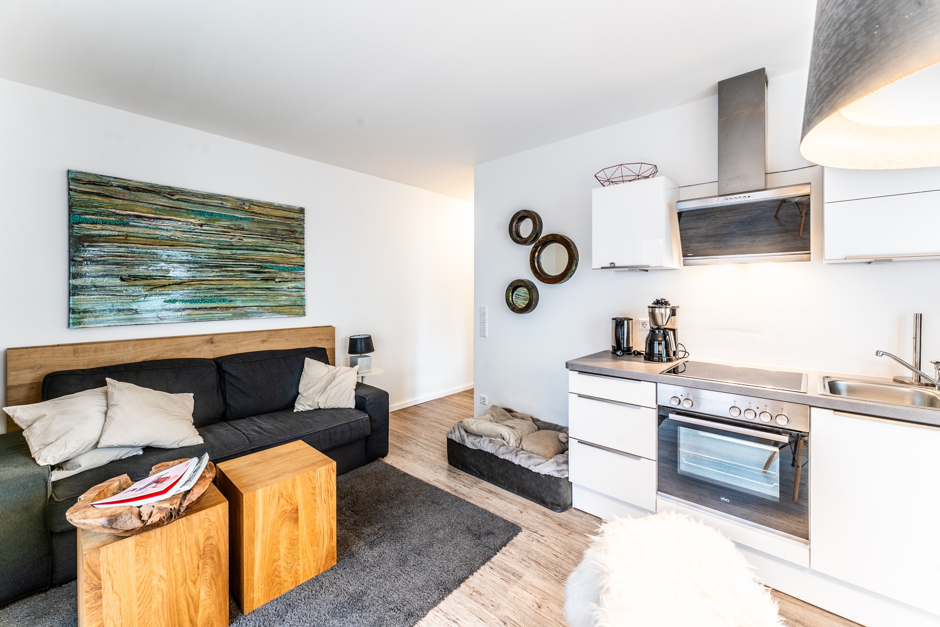Modernes Wohnzimmer mit schwarzem Sofa, Holztischen und offener Küche, Hundebett auf dem Boden.