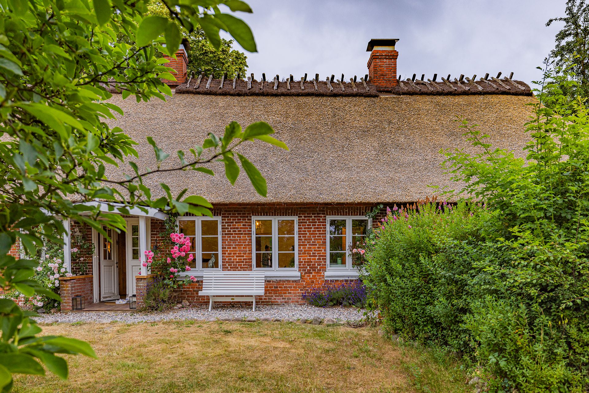 Backsteinhaus mit Reetdach, weißen Fenstern und Sitzbank, umgeben von Blumen und Büschen.
