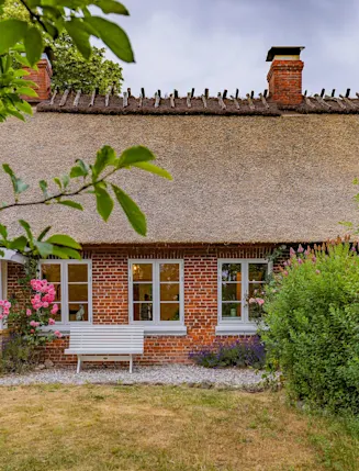 Backsteinhaus mit Reetdach, weißen Fenstern und Sitzbank, umgeben von Blumen und Büschen.