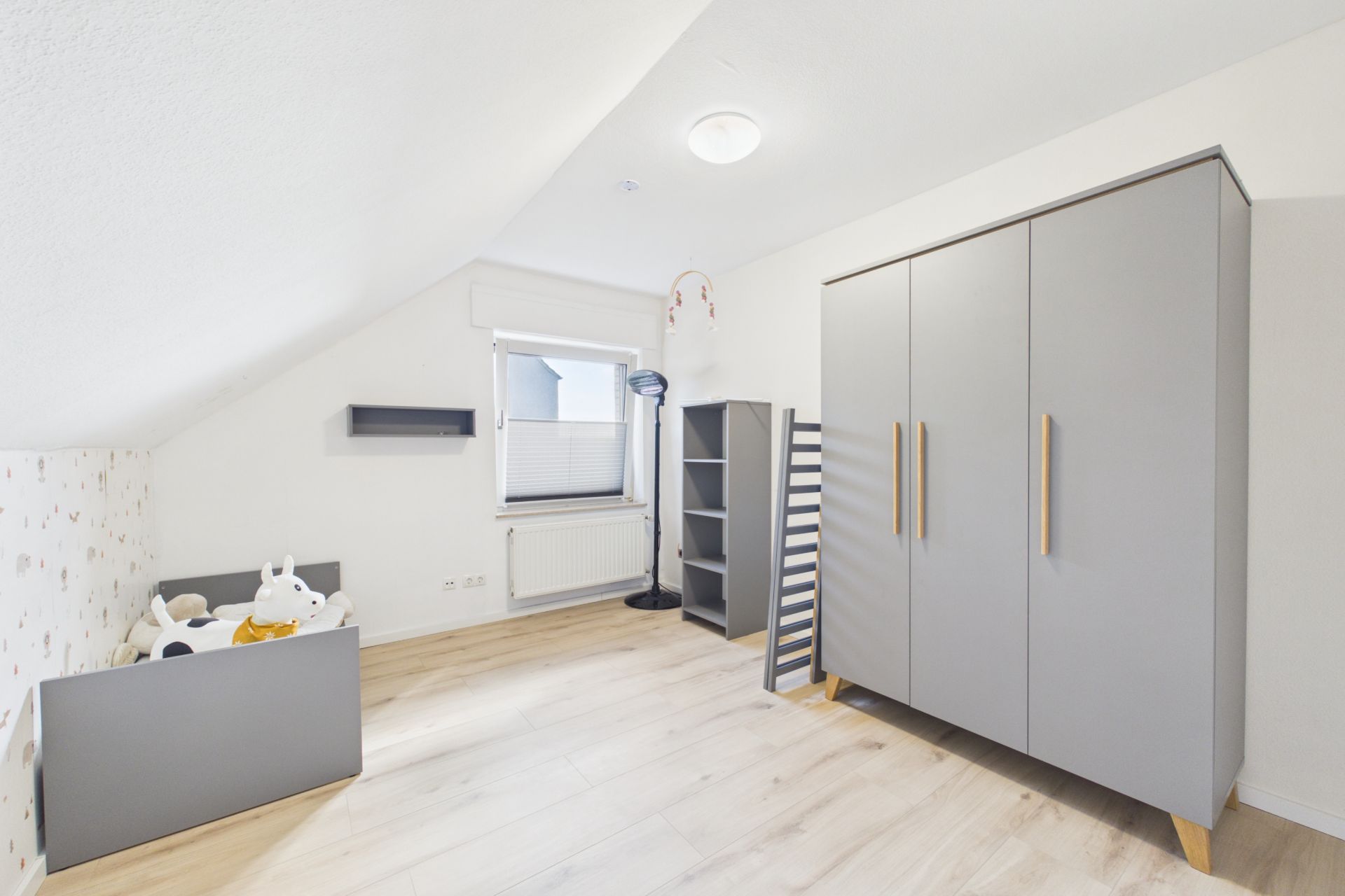Helles Kinderzimmer mit grauem Bett, Schrank, Regal und Fenster.