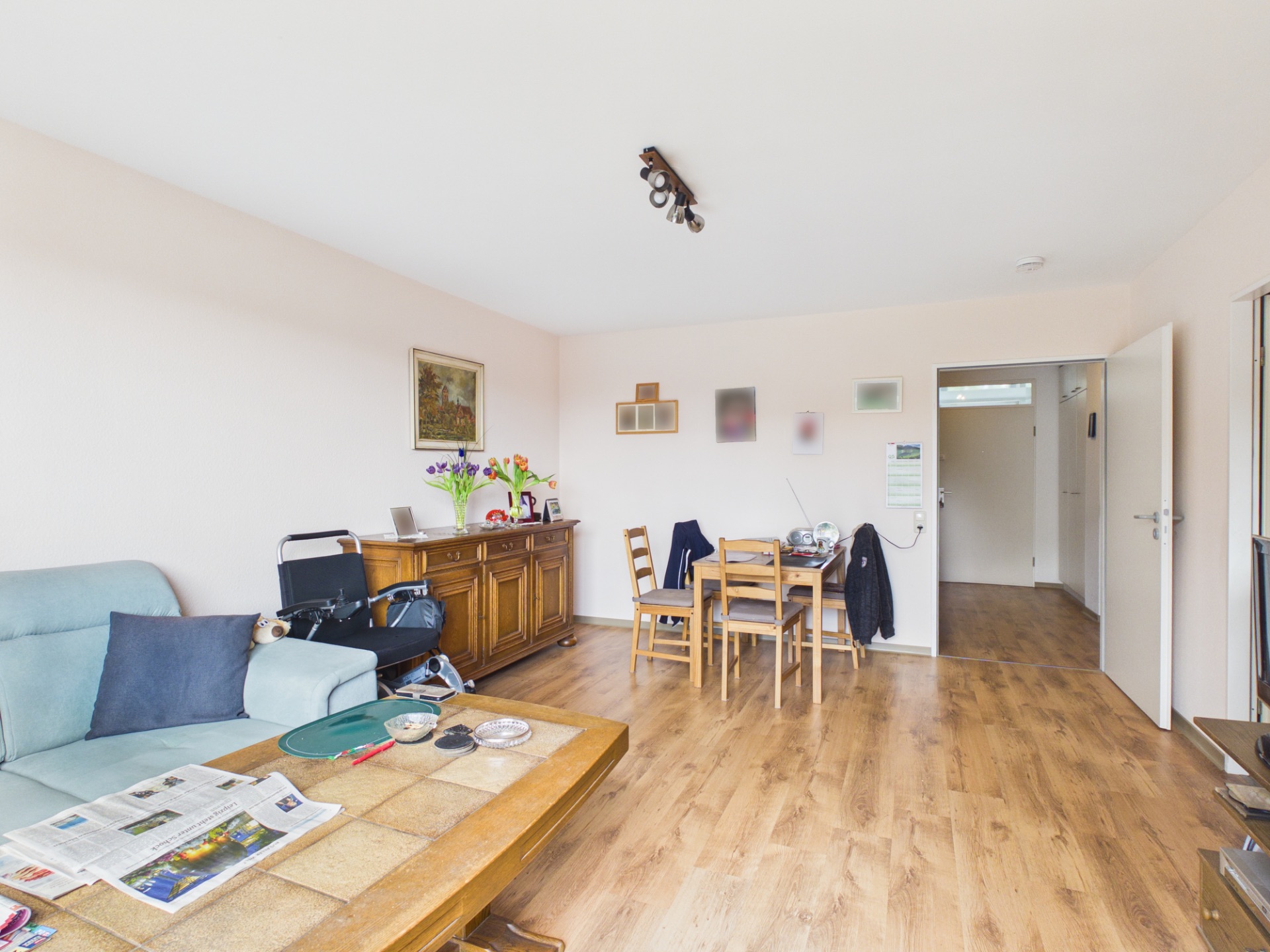Wohnzimmer mit Holzboden, Sofa, Esstisch und Kommode, Wanddekoration und offene Tür zum Flur.