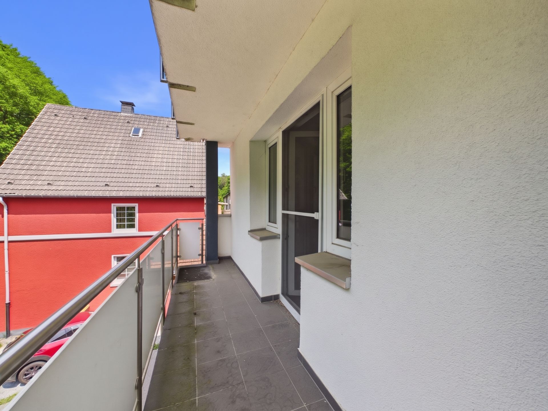Überdachter Balkon mit Glasgeländer und Blick auf ein rotes Wohnhaus.