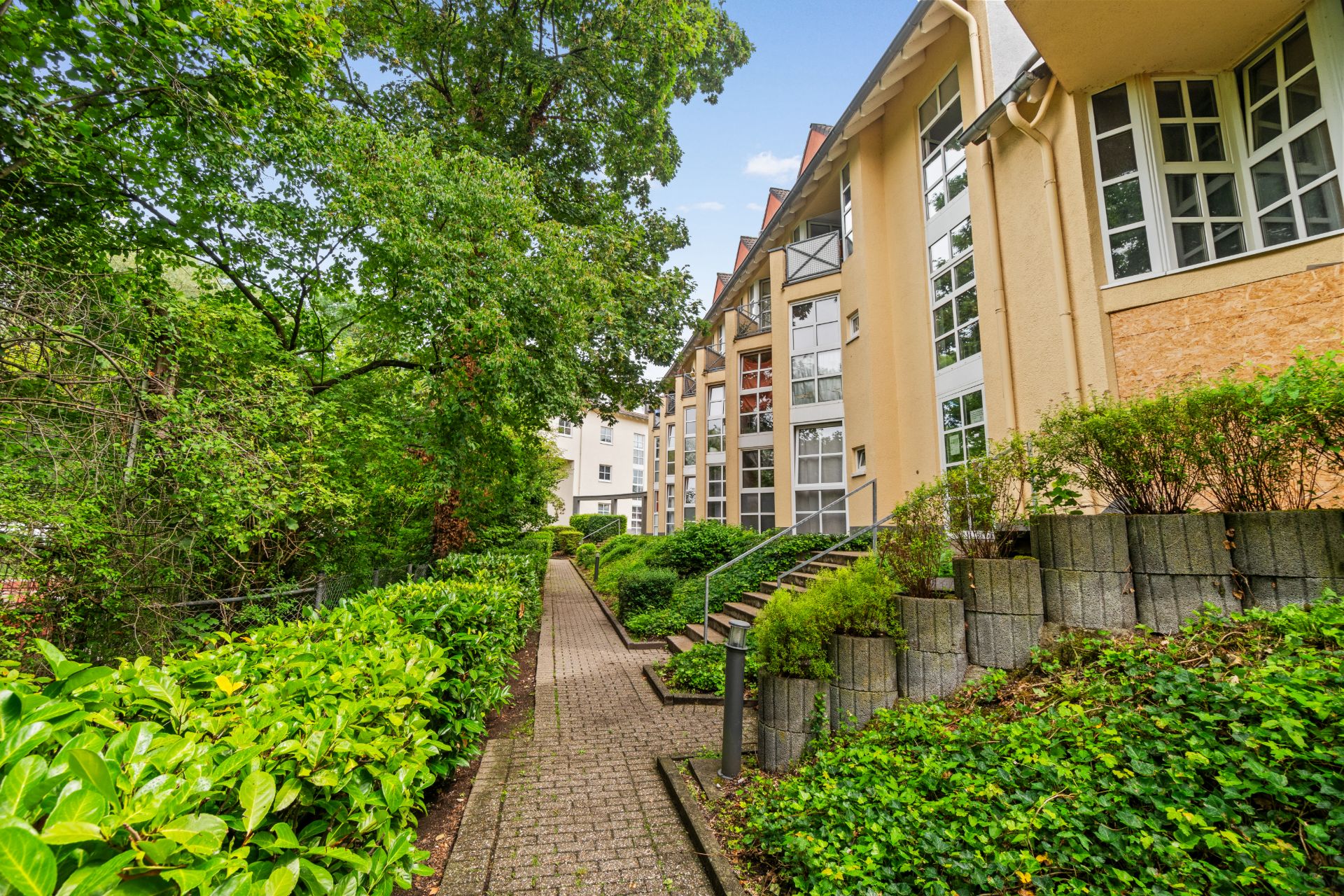 Gepflasterter Weg zwischen Wohngebäuden und grünen Büschen, Bäume im Hintergrund.