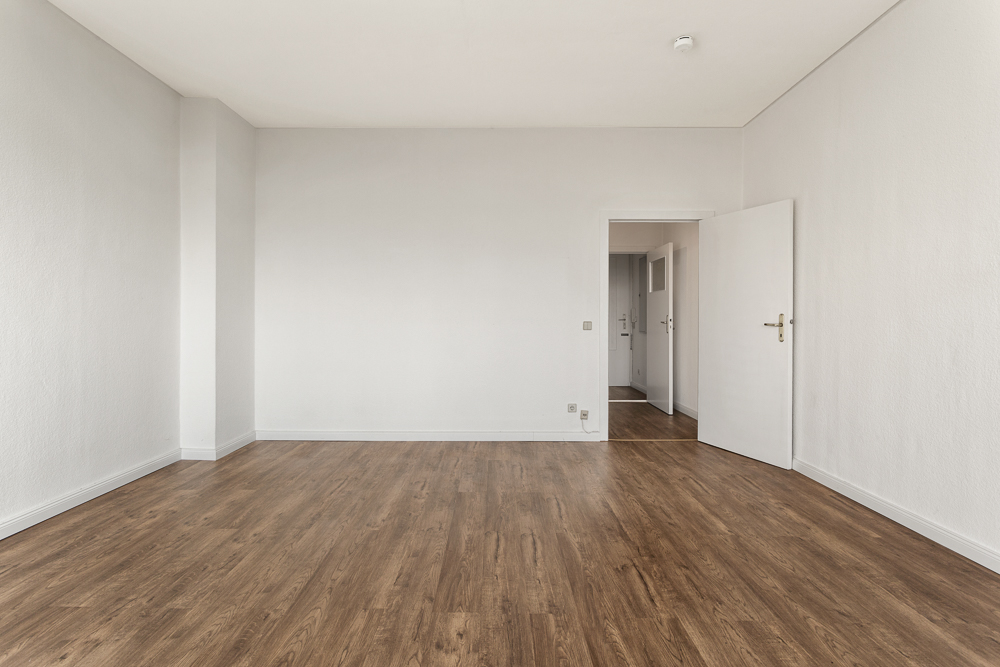 Leerer Raum mit weiß gestrichenen Wänden und braunem Holzboden, offene Tür im Hintergrund.