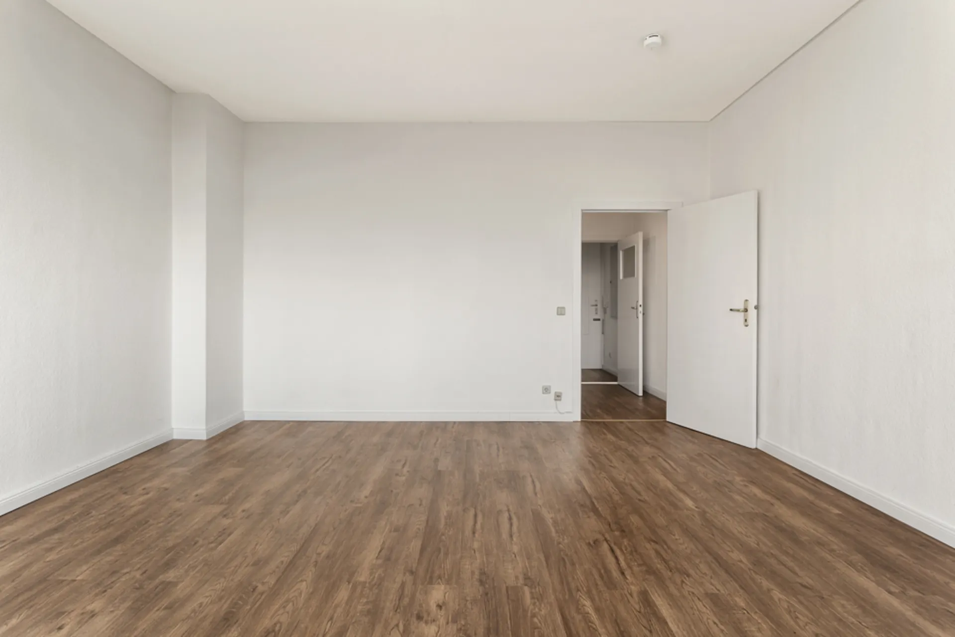 Leerer Raum mit weiß gestrichenen Wänden und braunem Holzboden, offene Tür im Hintergrund.