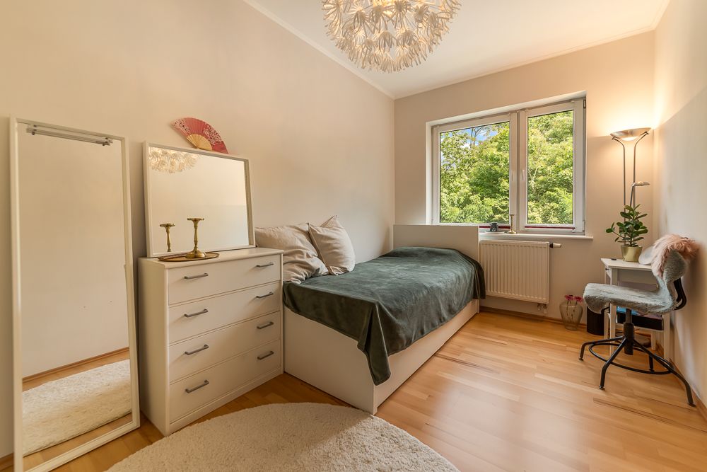 Helles Schlafzimmer mit Einzelbett, Kommode, Spiegel, Schreibtisch und großem Fenster mit Blick ins Grüne.