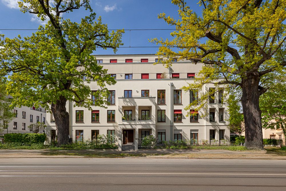 Mehrstöckiges Wohnhaus mit roten Fensterläden und großen Bäumen davor.