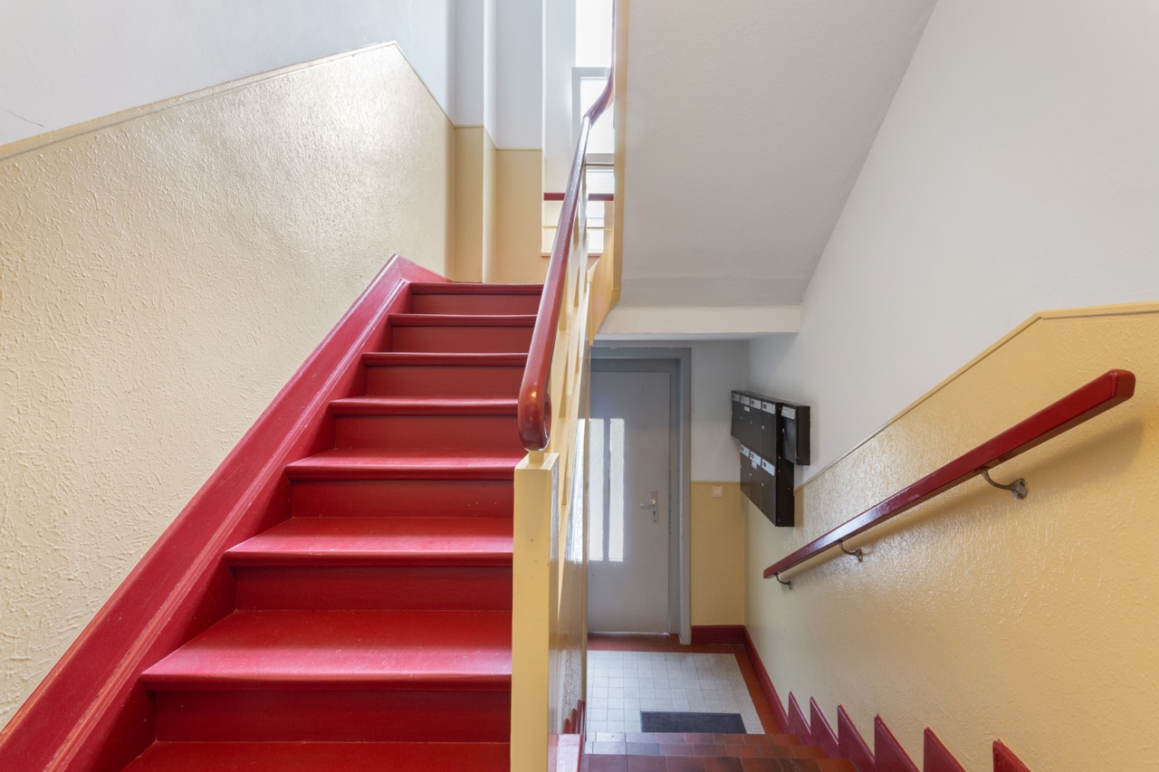 Treppenhaus mit roten Stufen und Geländer, gelbe Wände und Briefkästen an der Wand.