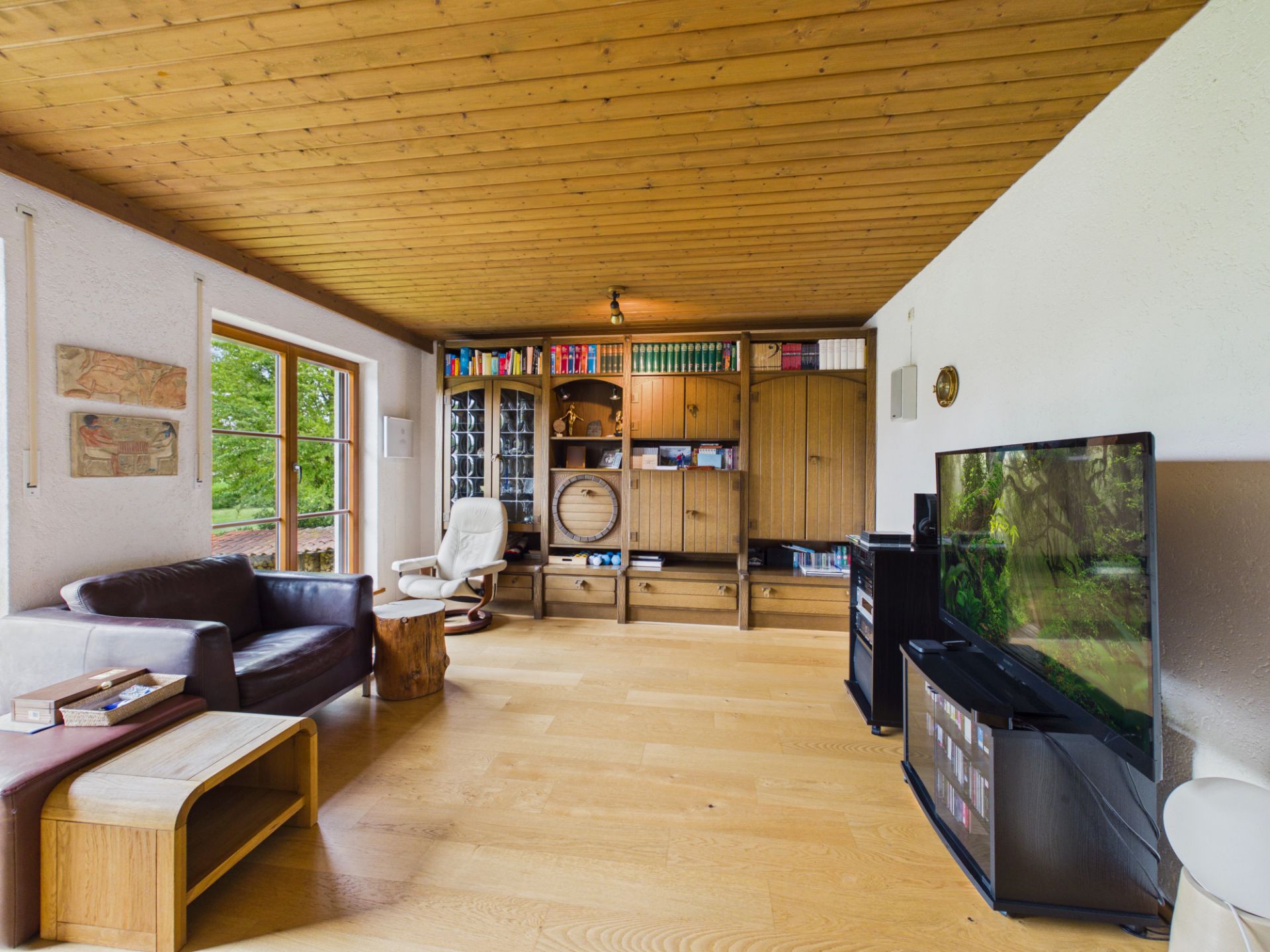 Helles Wohnzimmer mit Holzdecke, Bücherregal, Sofa, Sessel und großem Fernseher.