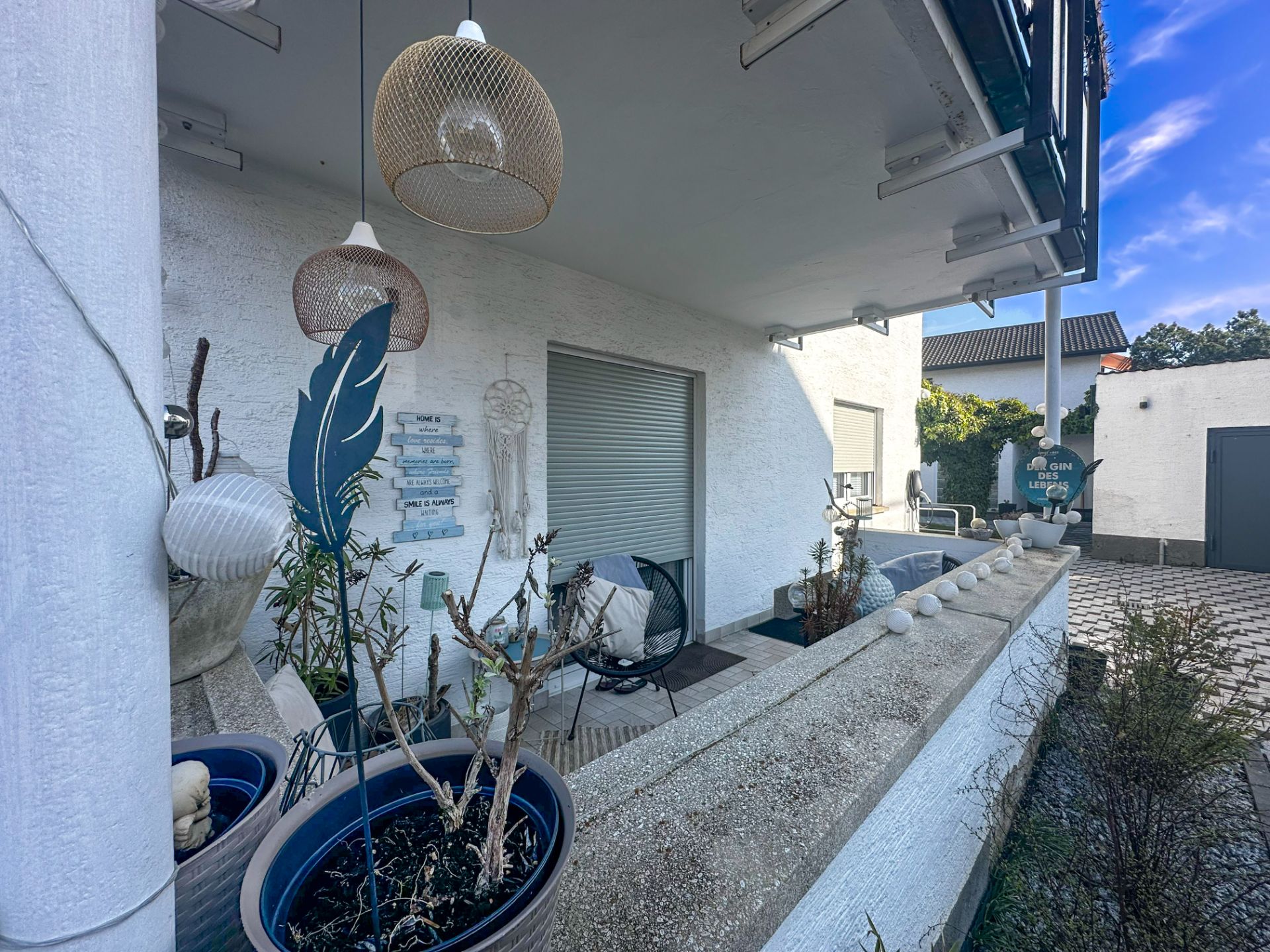 Überdachte Terrasse mit Pflanzen, Stühlen und Dekoration vor weißem Wohnhaus.