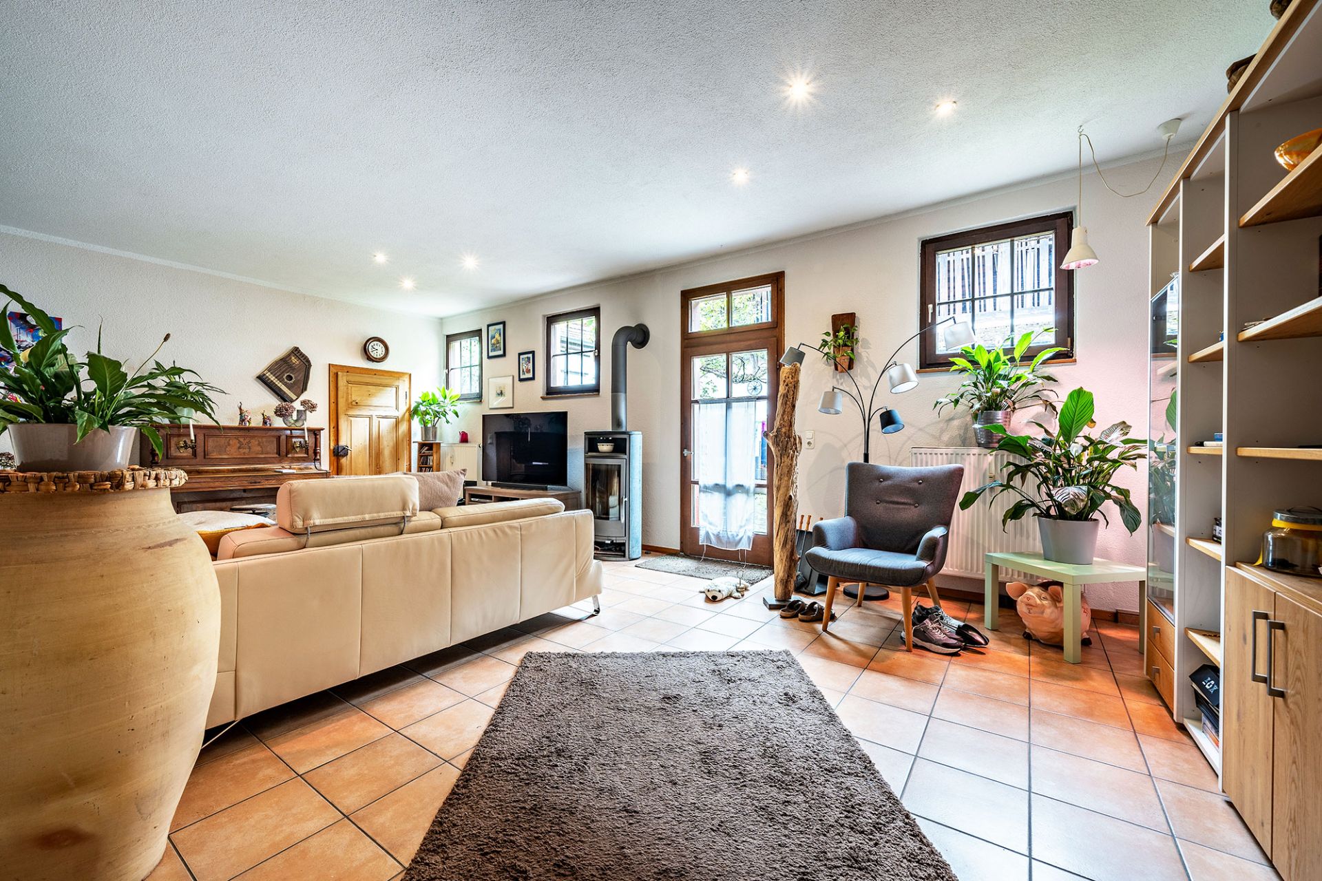 Helles Wohnzimmer mit Sofa, Sessel, Fernseher, Kaminofen, Pflanzen und Regalen.