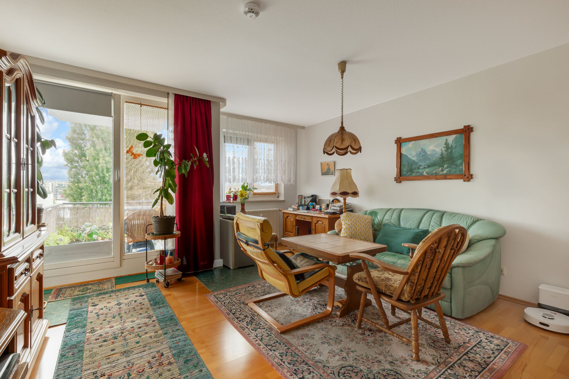 Wohnzimmer mit grünem Sofa, Holztisch, Teppichen, großen Fenstern und Zugang zum Balkon.