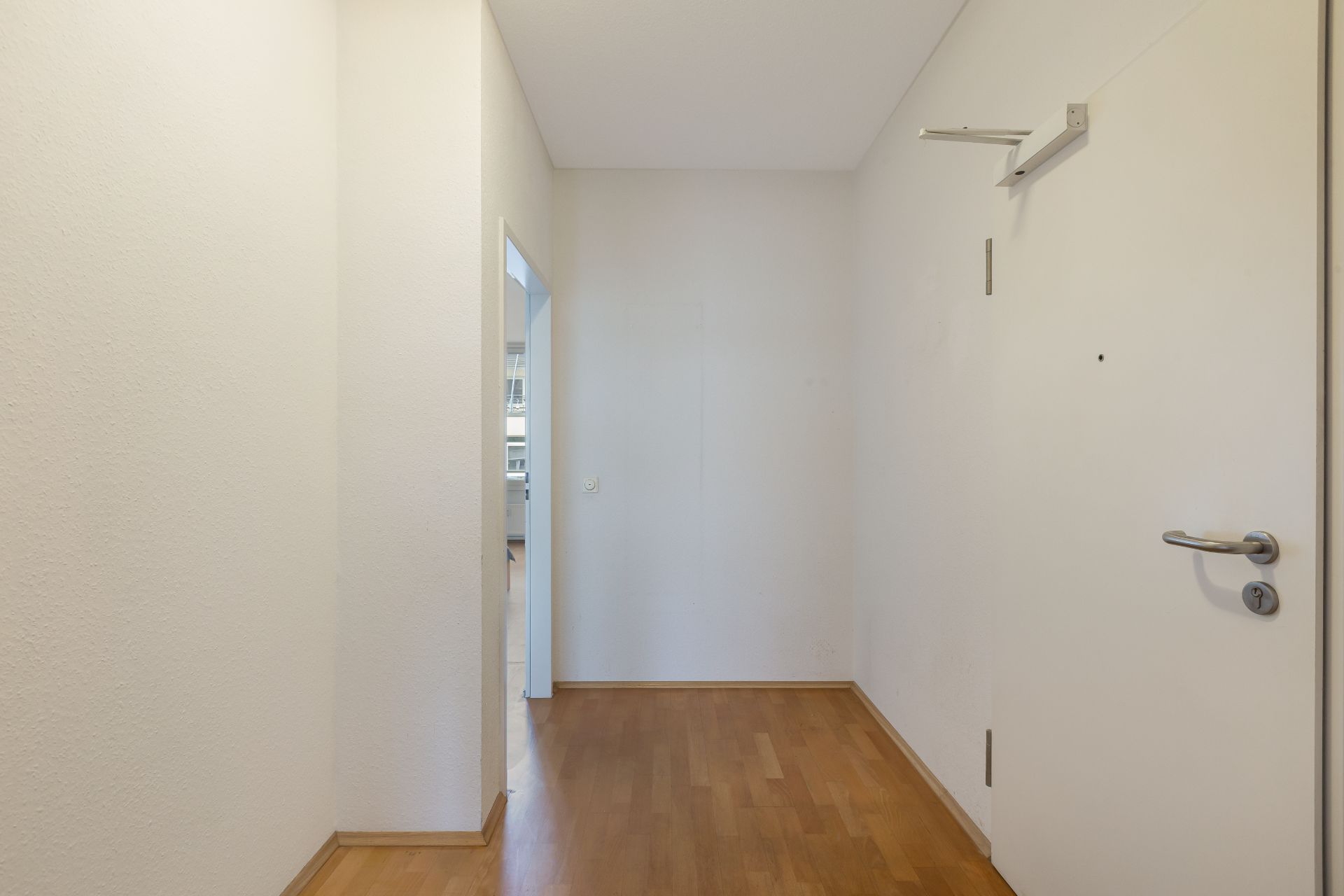 Heller Flur mit Holzboden, weißer Wand und offener Tür zu einem angrenzenden Raum.