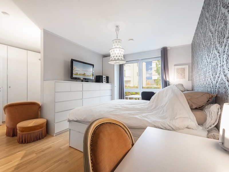 Helles Schlafzimmer mit Bett, Kommode, Fernseher, Stuhl und Fenster mit Blick nach draußen.