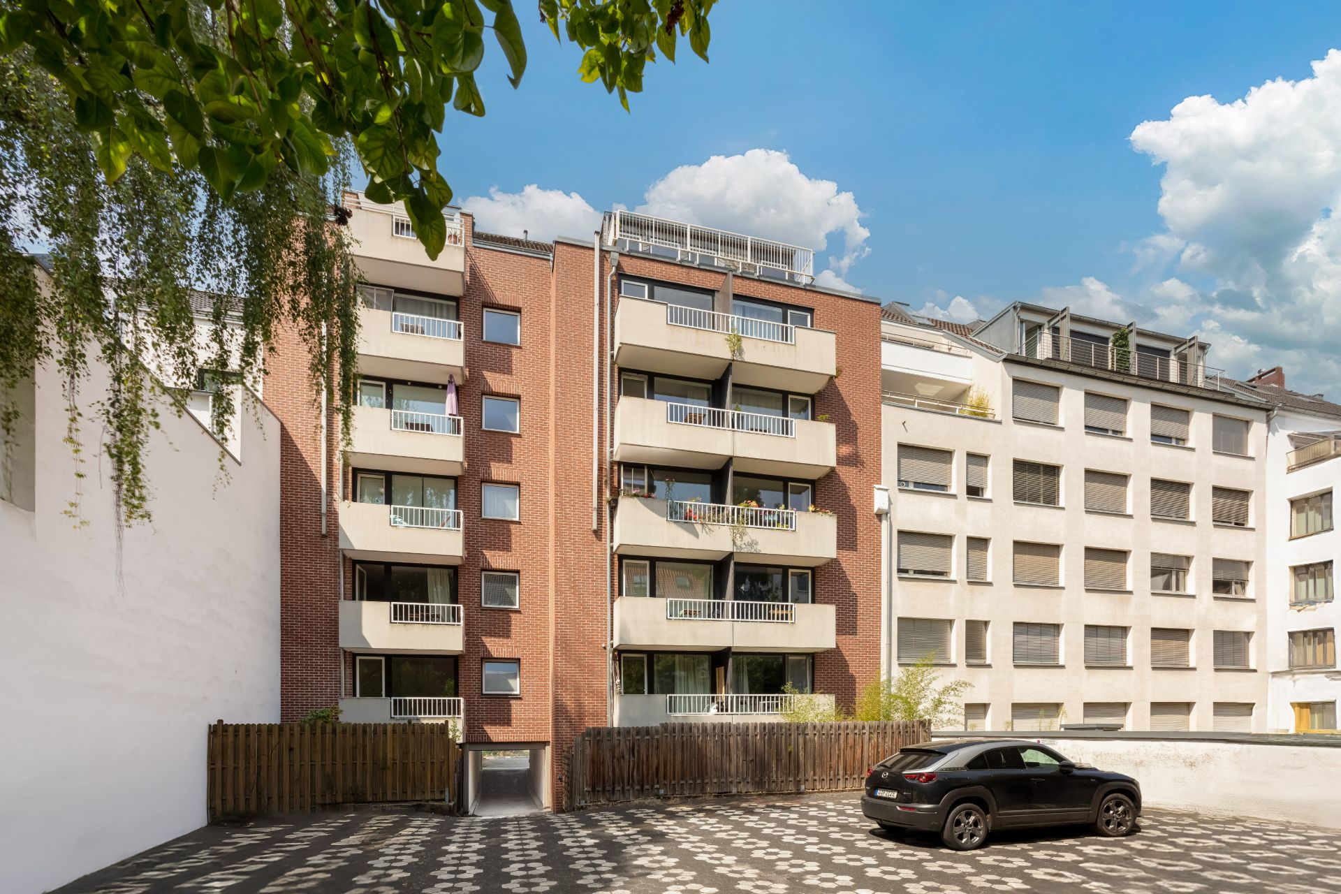 Mehrfamilienhaus mit Balkonen, Parkplatz im Vordergrund, ein Auto parkt auf gemustertem Boden.