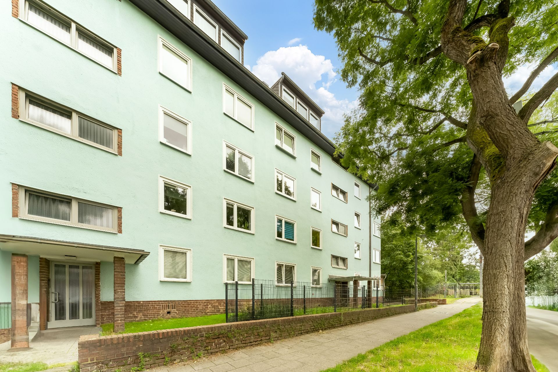 Mehrstöckiges Wohnhaus mit vielen Fenstern, Eingängen und einem großen Baum davor.