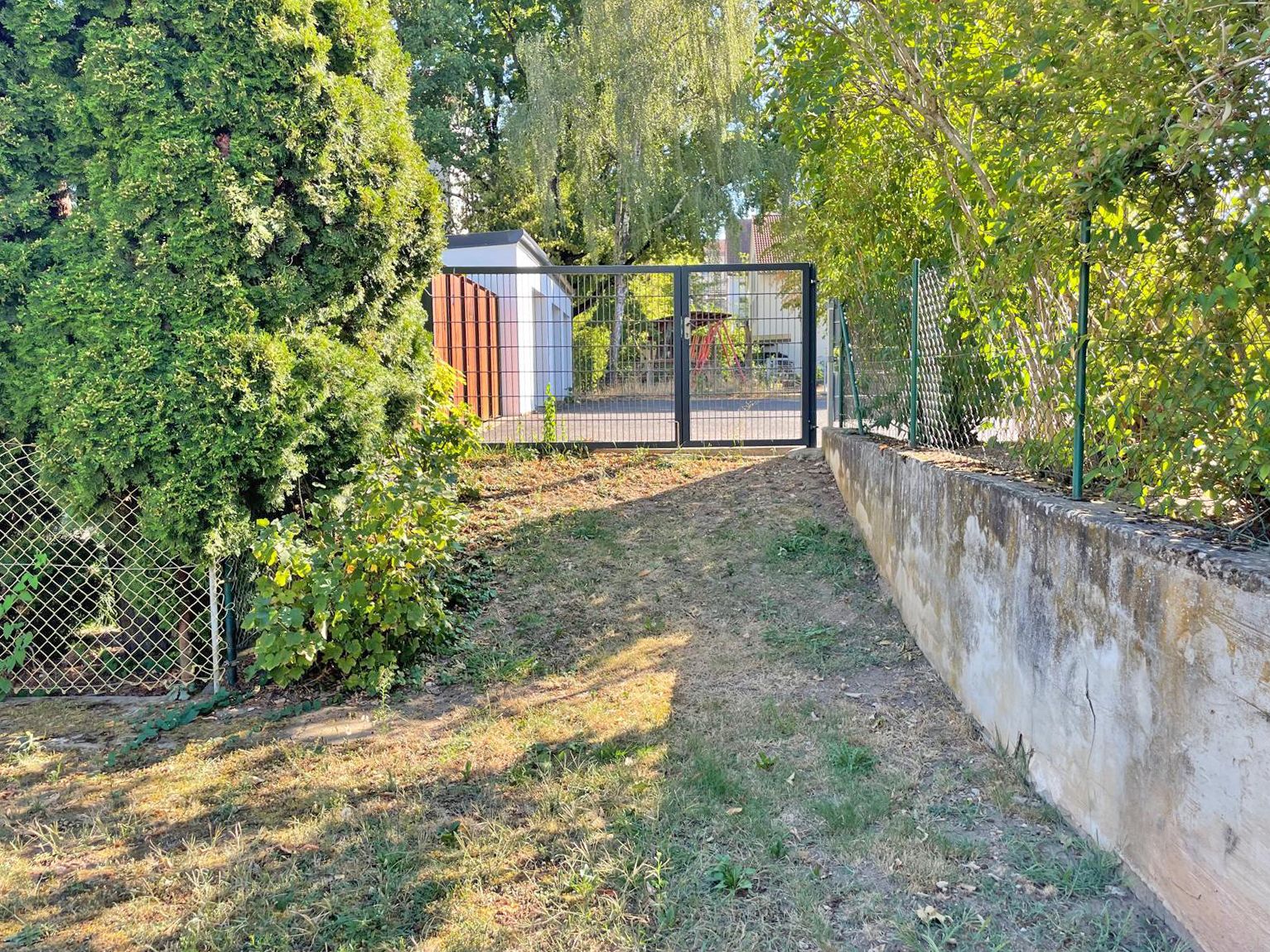 Gartenweg mit Metallzaun und Tor, umgeben von Bäumen und Sträuchern.