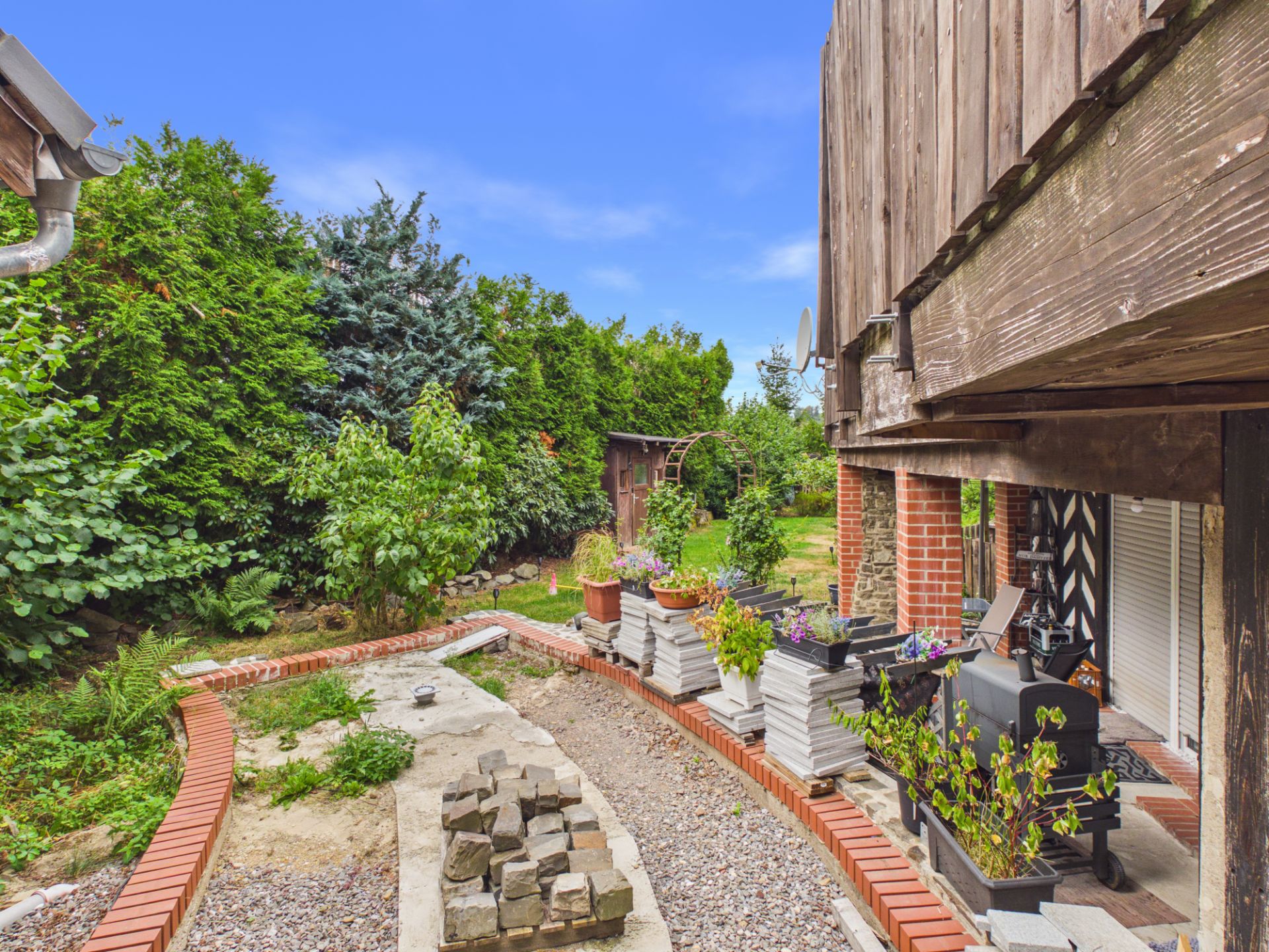 Garten mit Pflanzen, Bäumen, Steinweg und einer Terrasse neben einem Wohnhaus.