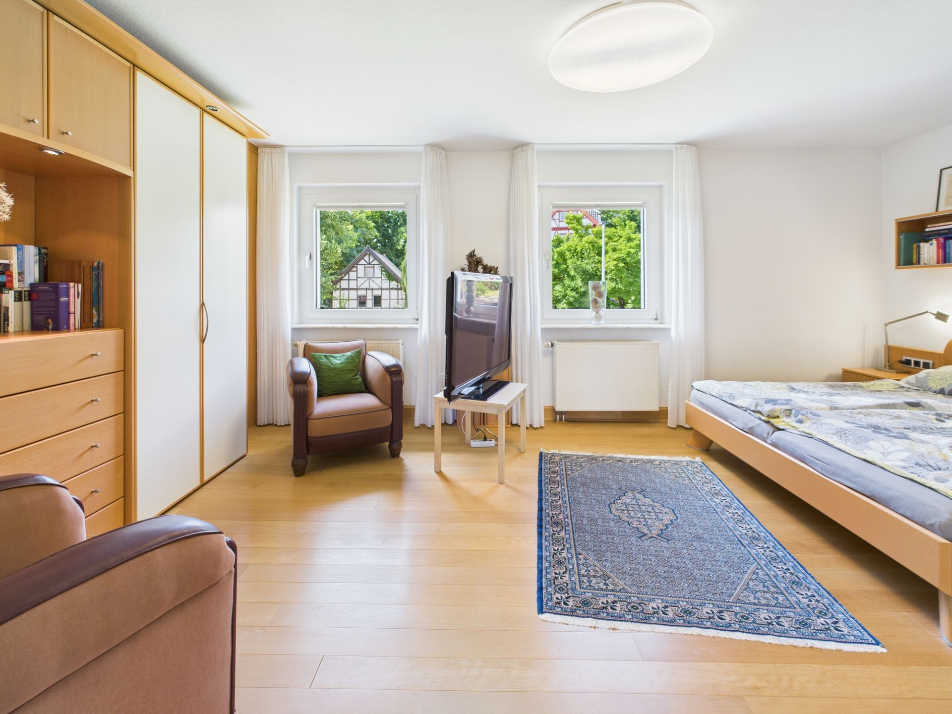 Helles Schlafzimmer mit Doppelbett, Teppich, Sessel, Kleiderschrank, Fernseher und zwei Fenstern.