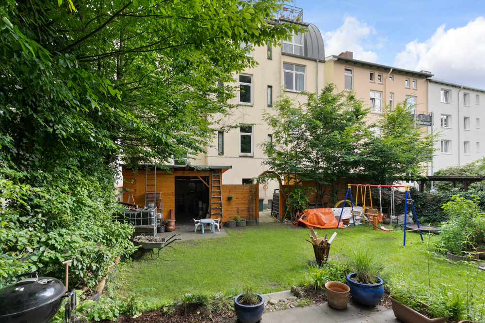Grüner Hinterhof mit Rasen, Schaukel, Gartenhaus und Blumen in Töpfen.