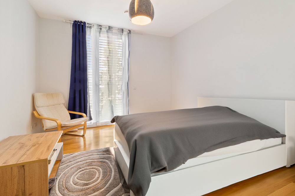 Helles Schlafzimmer mit Bett, Sessel, Teppich, Kommode und großen Fenstern mit Vorhängen.