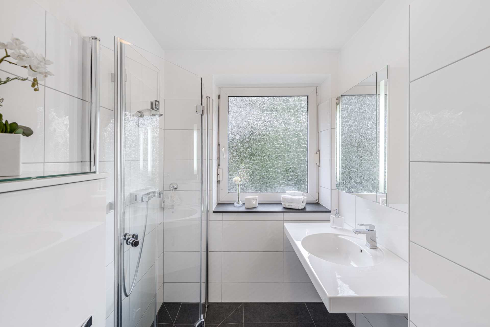 Modernes, weiß gefliestes Badezimmer mit Dusche, Waschbecken und Fenster mit Milchglas.