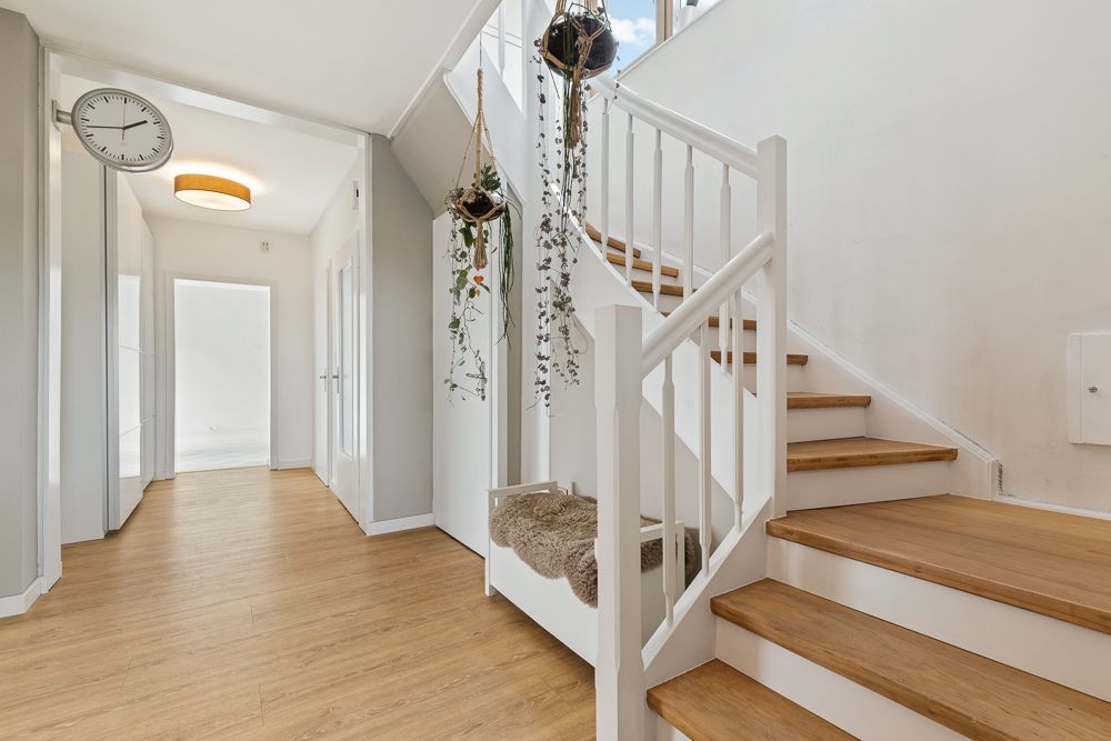 Heller Flur mit Holztreppe, hängenden Pflanzen und Wanduhr, Parkettboden.
