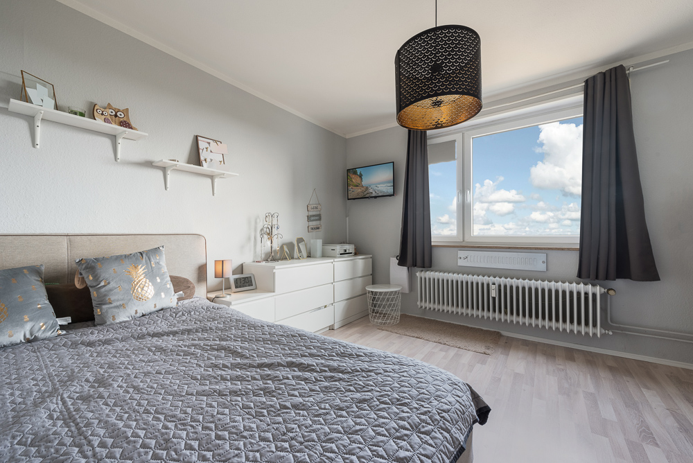 Modernes Schlafzimmer mit grauer Bettdecke, großem Fenster und weißer Kommode.