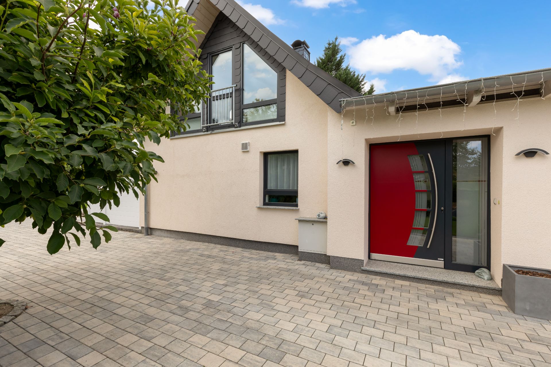 Modernes Einfamilienhaus mit roter Haustür, gepflastertem Hof und Baum im Vordergrund.