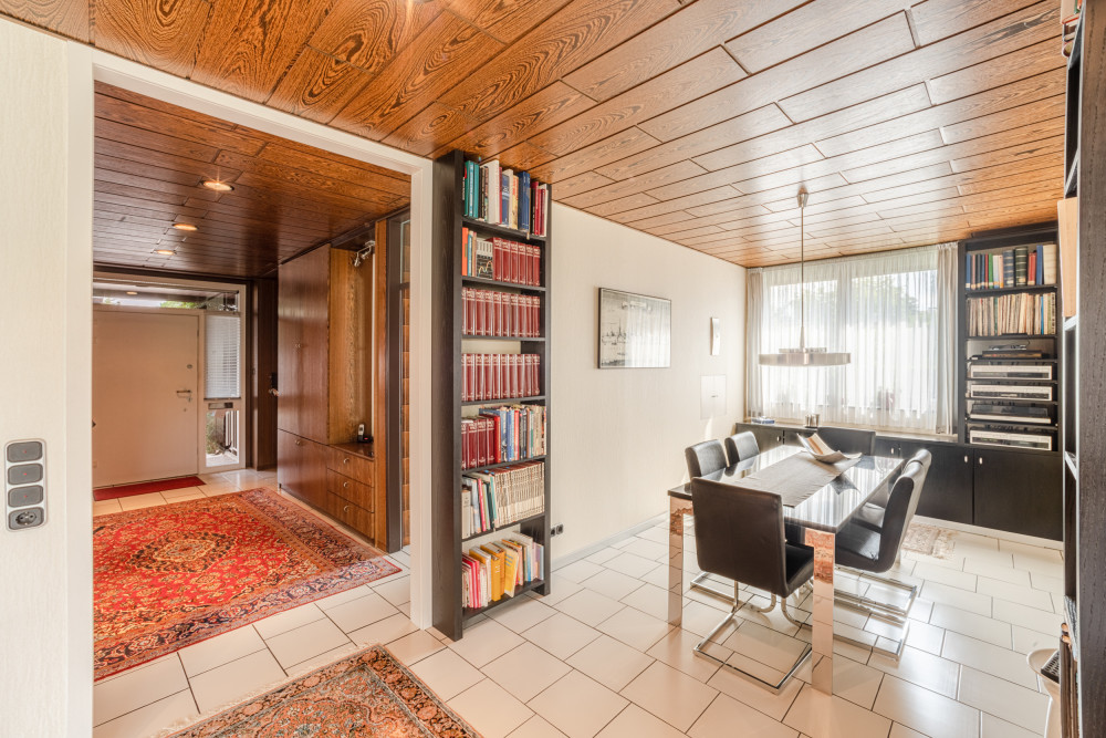 Esszimmer mit Glastisch, vier Stühlen, Bücherregal und großen Fenstern. Teppiche auf weißen Fliesen.