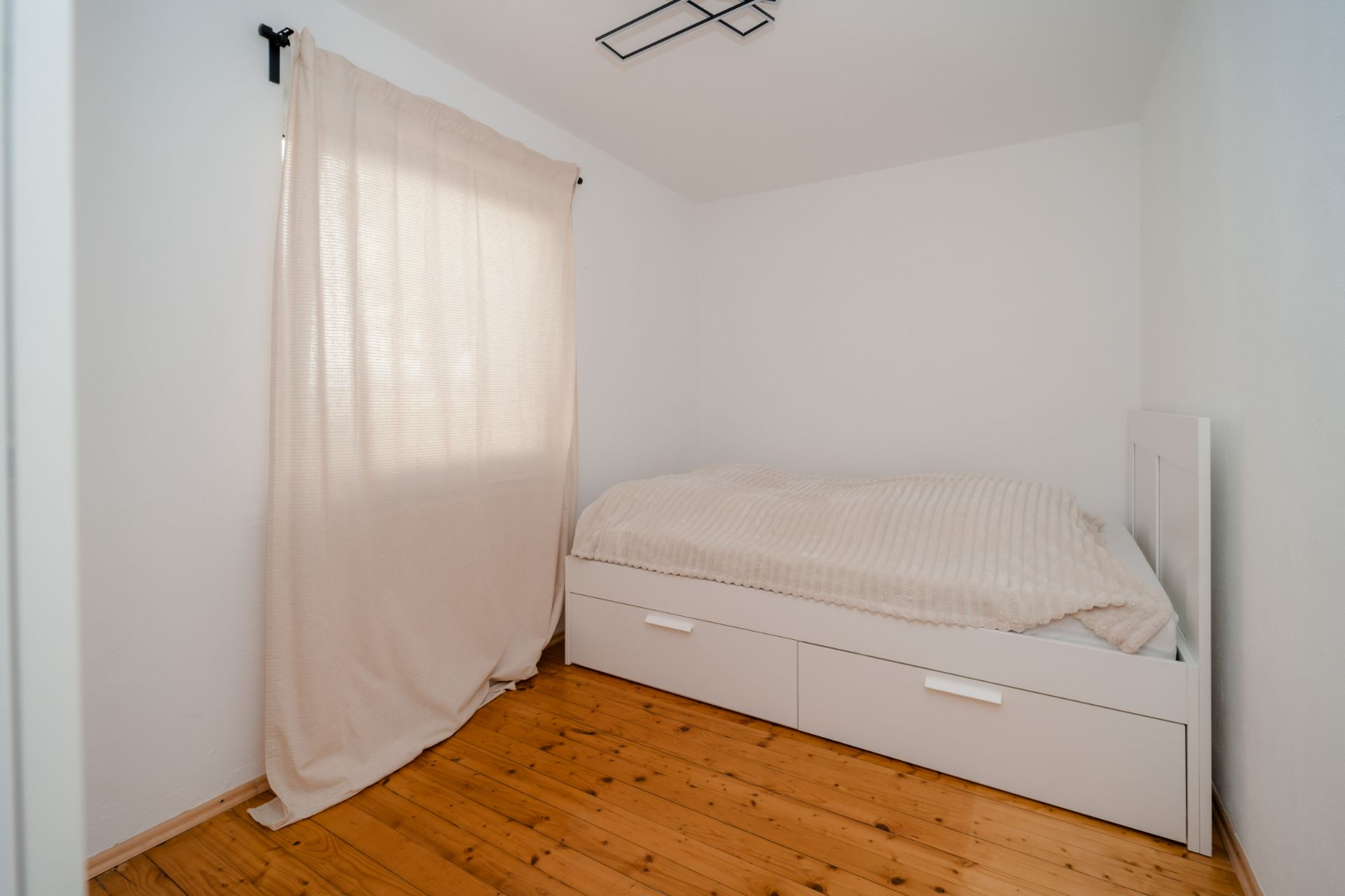 Kleines Schlafzimmer mit Einzelbett, weißer Bettwäsche, Holzboden und hellem Vorhang am Fenster.