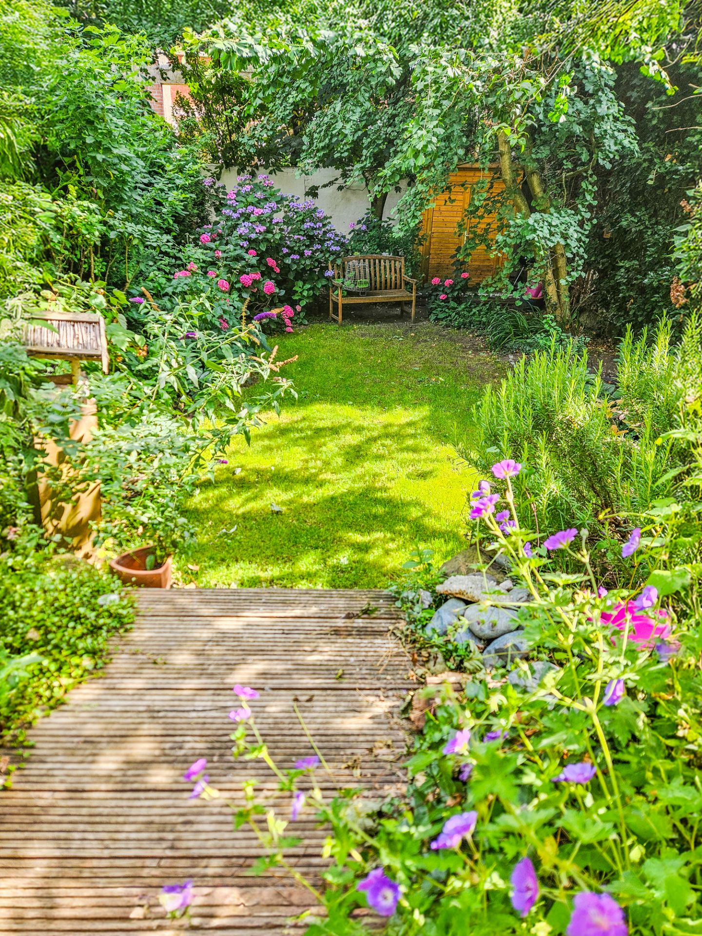 Grüner Garten mit Holzboden, bunten Blumen, Rasen und Holzbank im Hintergrund.
